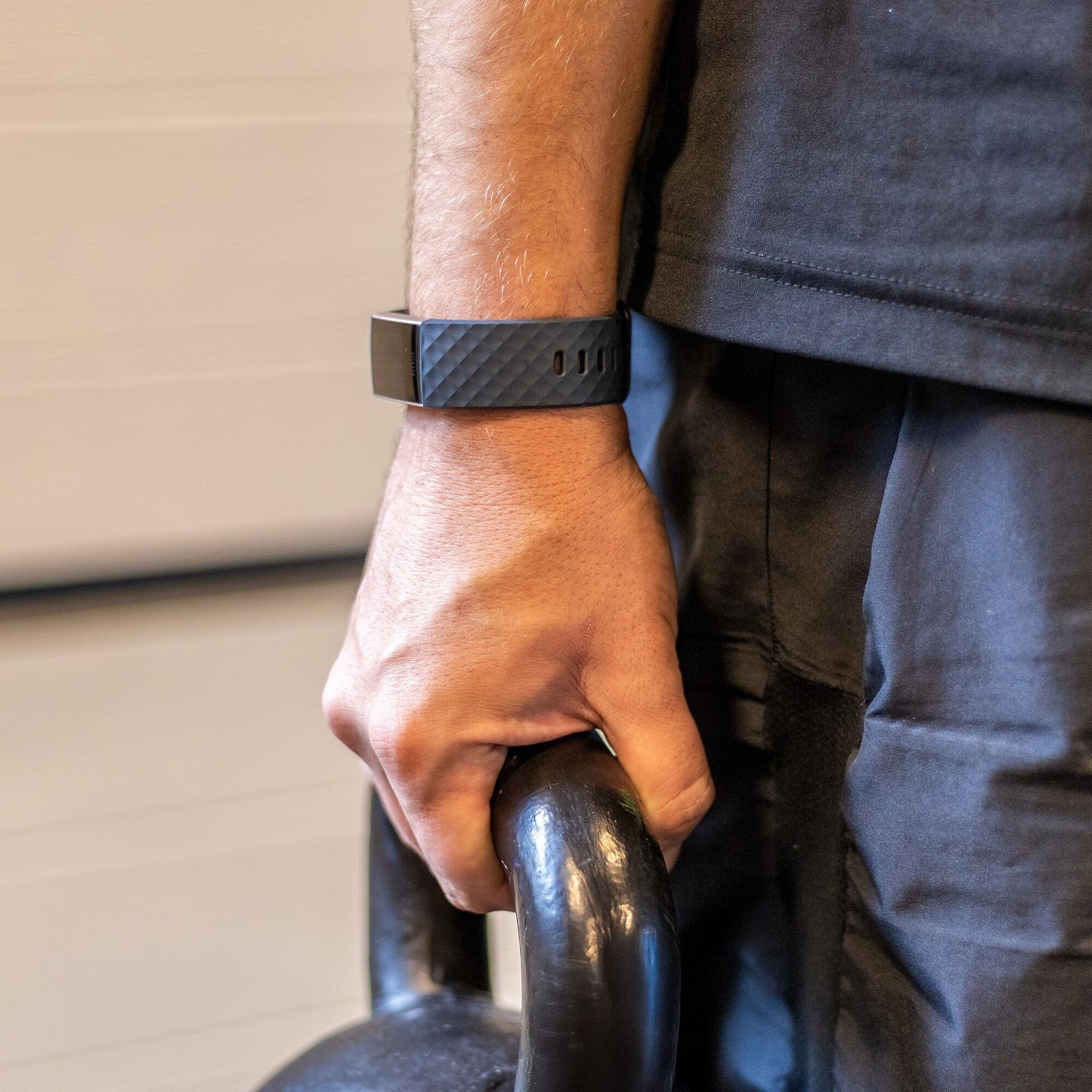 Eine Person hält eine schwarze Kettlebell. Sie trägt einen schwarzen Fitnesstracker am Handgelenk und ein schwarzes Hemd.