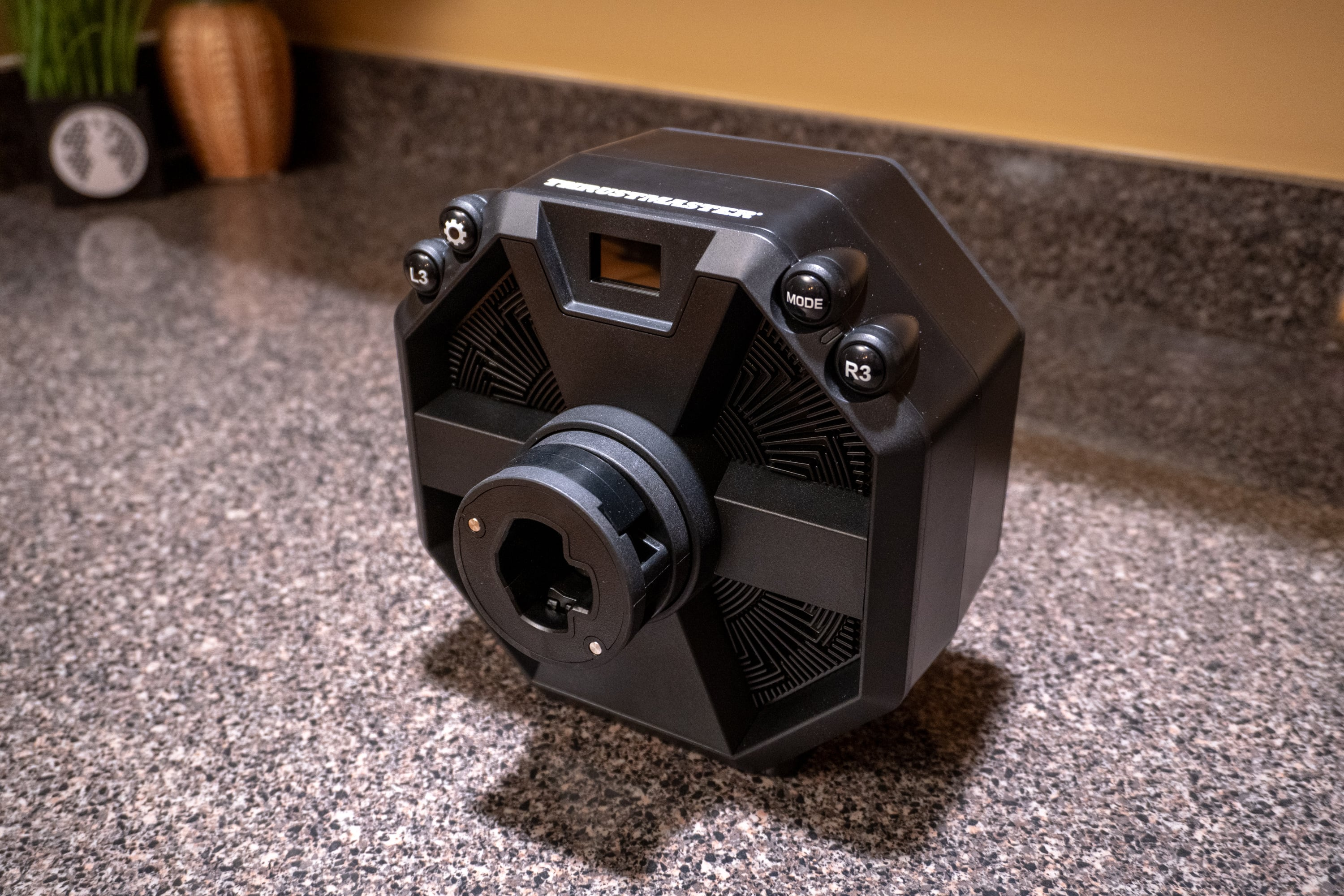 Ein schwarzes Thrustmaster-Rennrad liegt auf einer gesprenkelten Arbeitsplatte. Knöpfe mit der Aufschrift L3, Mode und R3 sind sichtbar.
