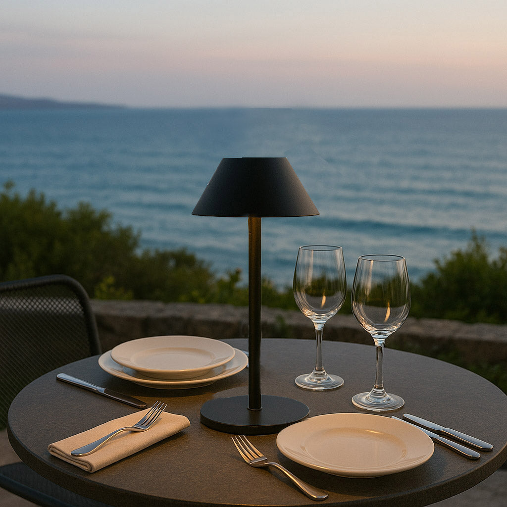 Tisch mit Lampe, Tellern, Weingläsern und Besteck mit Blick auf das Meer in der Abenddämmerung. Details: Serviette und Stuhl.