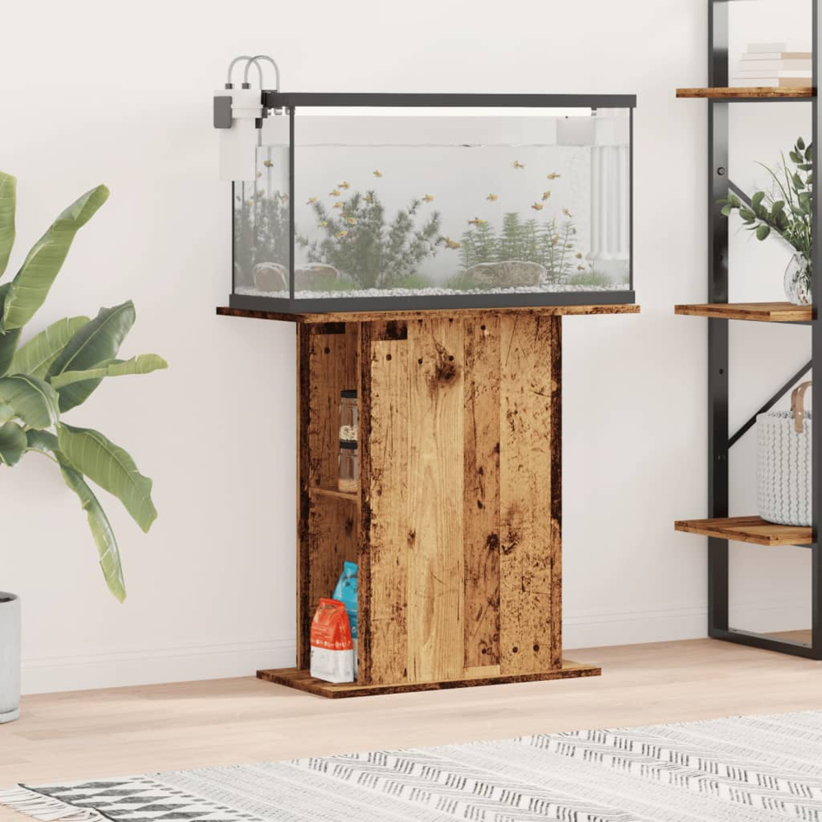 Ein Aquarium mit Fischen steht auf einem Holzständer. Pflanzen und ein Regal sind im Hintergrund. Ein Teppich und eine Topfpflanze sind davor.