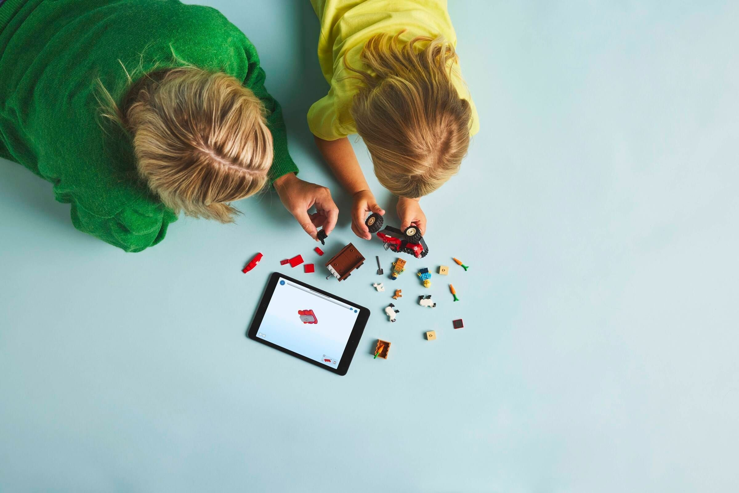 Zwei Kinder bauen Spielzeugteile auf einer blauen Oberfläche zusammen. Es gibt ein Tablet mit Anweisungen und verstreuten Spielzeugteilen.