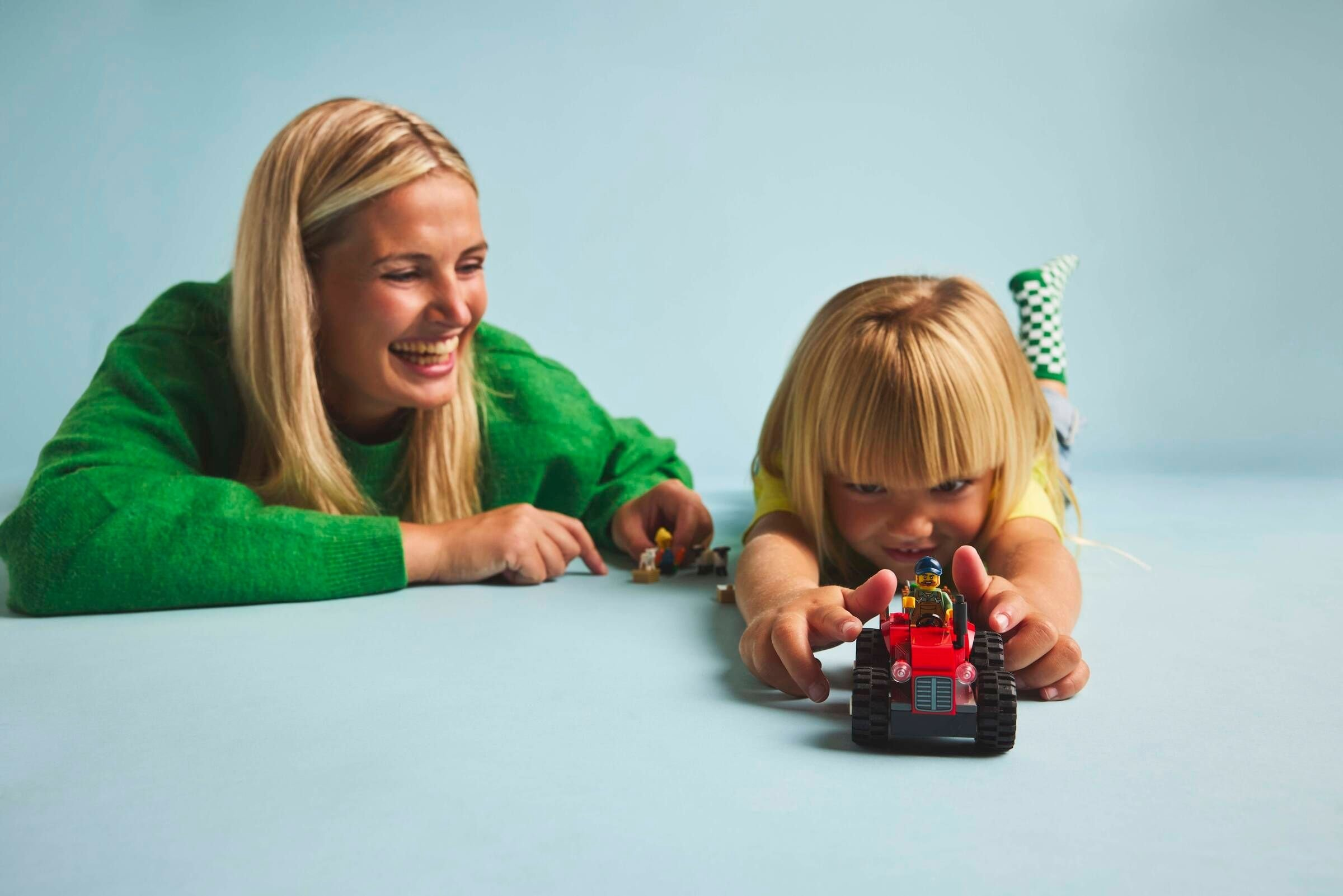 Eine Frau und ein Kind spielen mit Lego. Das Kind hält einen roten Traktor. Die Frau lächelt.