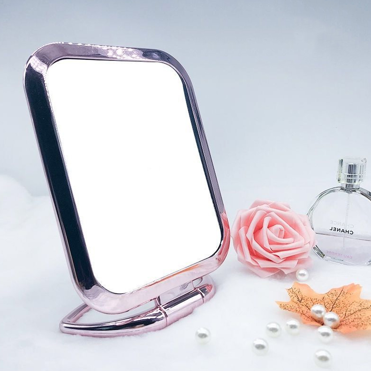 Ein roségoldener Spiegel steht neben einer rosa Rose, einer Parfümflasche, Perlen und einem Blatt auf weißem Untergrund.