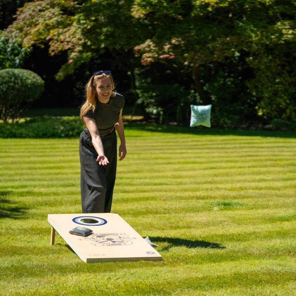 Eine Frau wirft einen Beanbag auf ein Cornhole-Board in einem grasbewachsenen Garten. Ein Beanbag ist in der Luft. Bäume und Büsche sind im Hintergrund.