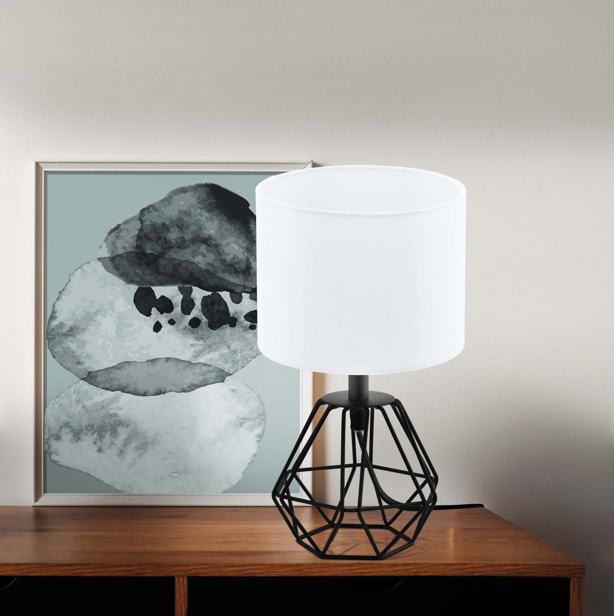 Una lampada con paralume bianco e base geometrica nera su superficie in legno. Un'opera d'arte incorniciata sullo sfondo.
