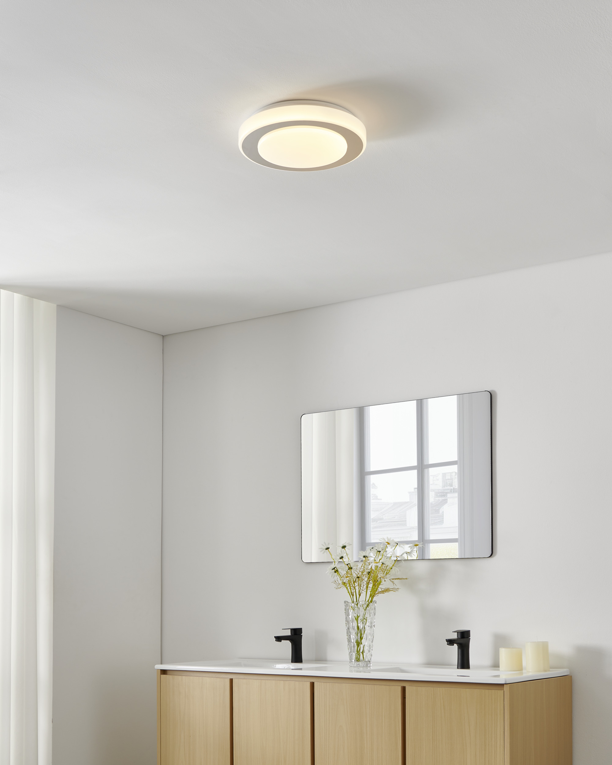 Badezimmer mit Deckenleuchte, Spiegel, Waschbecken, Blumen und Kerzen. Unterschrank aus hellem Holz.