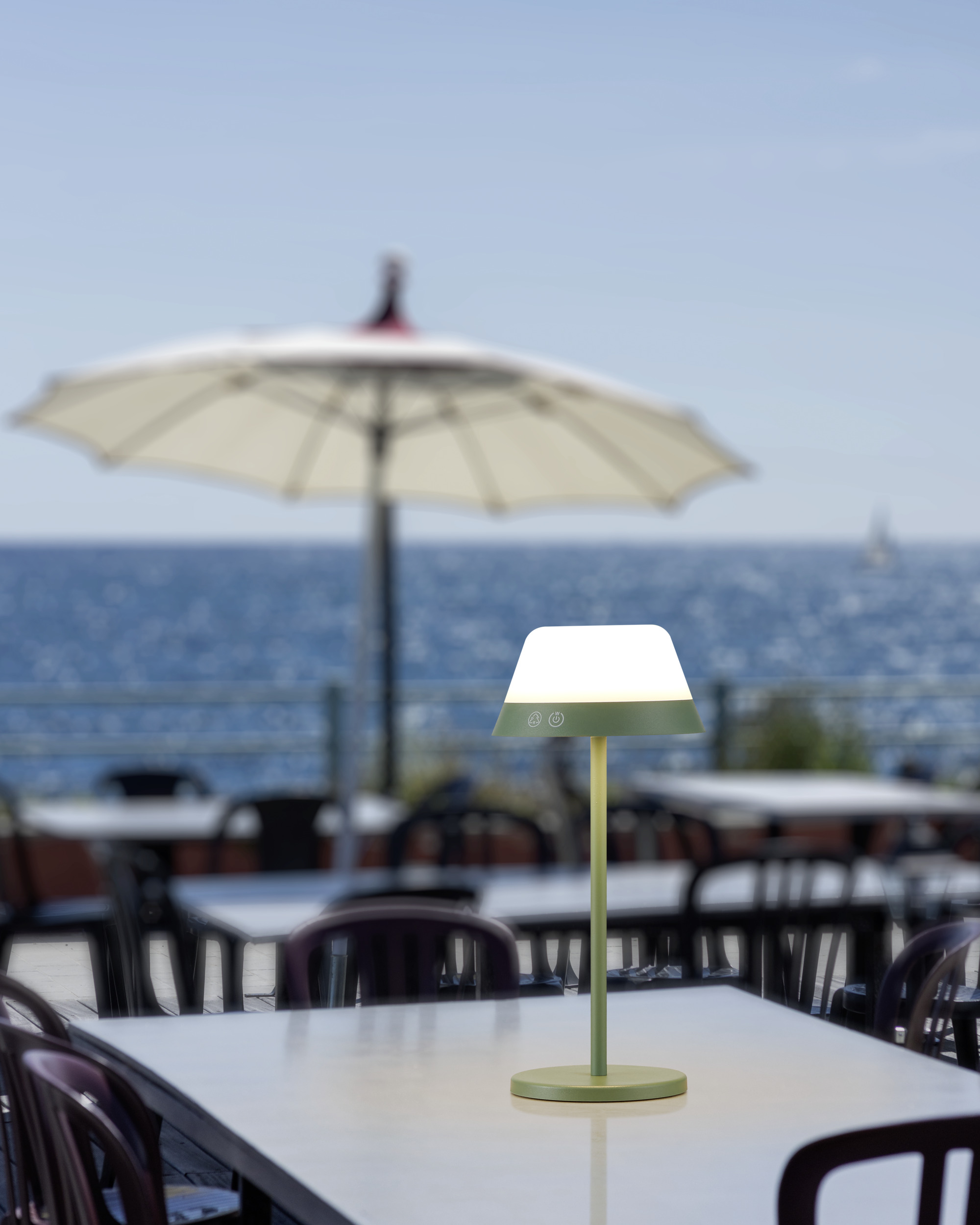 Eine grüne Lampe mit weißem Lampenschirm auf einem weißen Tisch. Im Hintergrund ein Strandrestaurant.