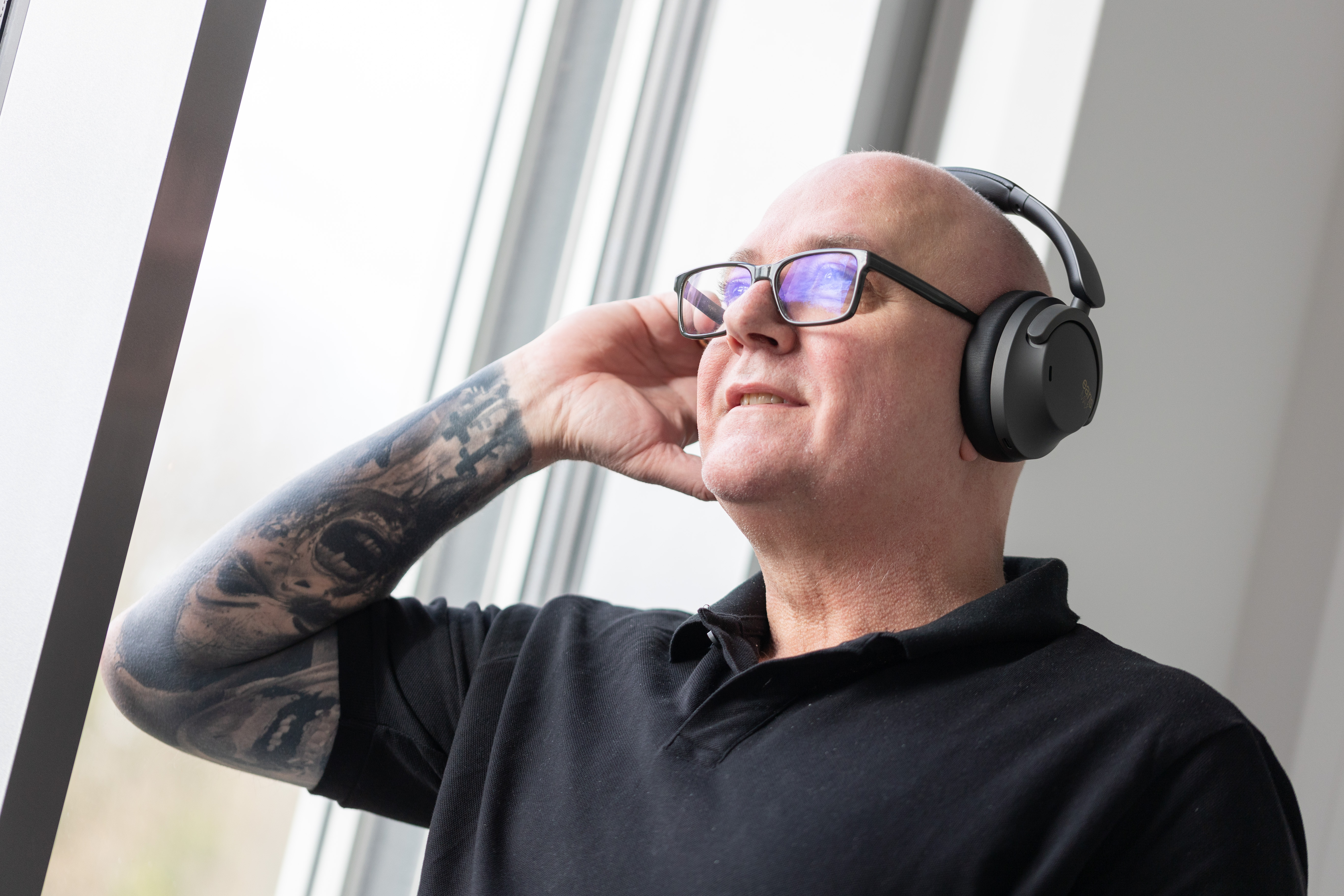 Glatzköpfiger Mann mit Brille und Kopfhörern. Tätowierung am Arm. Trägt ein schwarzes Poloshirt. An einem Fenster lehnend.