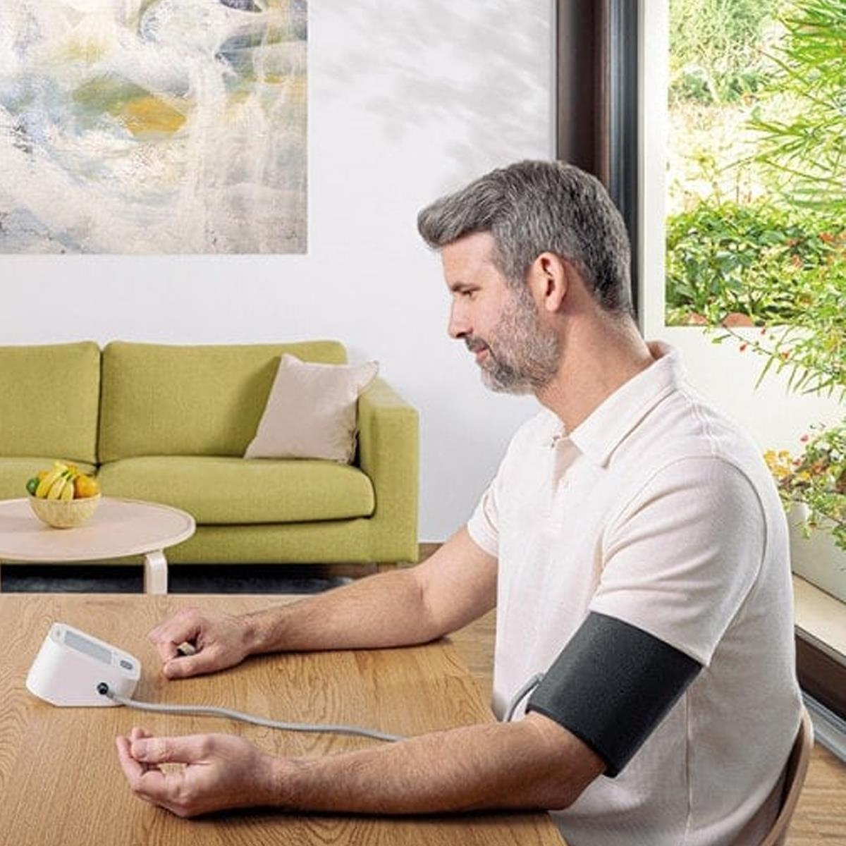 Ein Mann misst seinen Blutdruck mit einem Gerät. Eine Manschette ist an seinem Arm. Sofa und Fenster im Hintergrund.