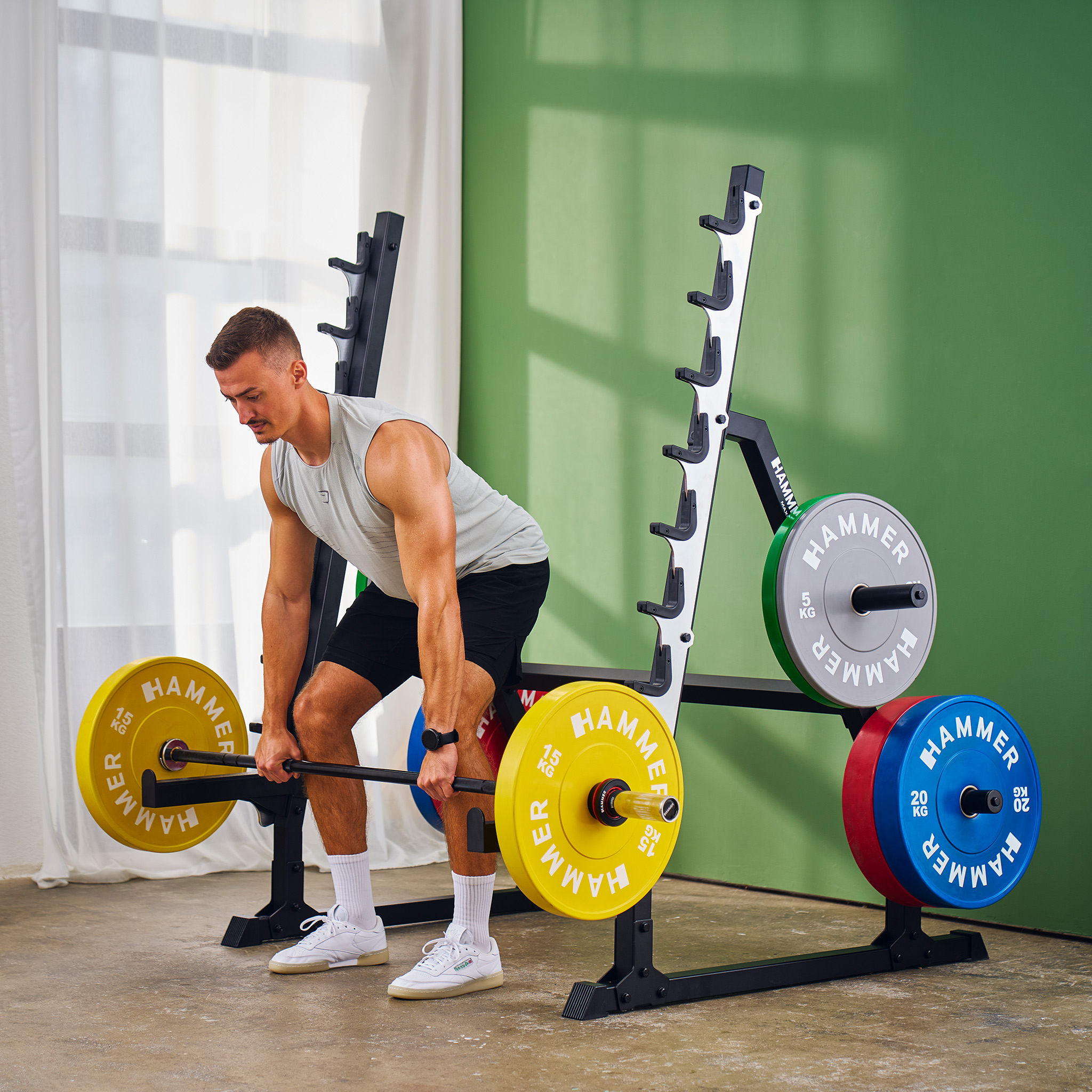 Mann, der eine Langhantel mit gelben, grauen und blauen Scheiben in einem Fitnessstudio hebt. Hantelständer und grüne Wand im Hintergrund.