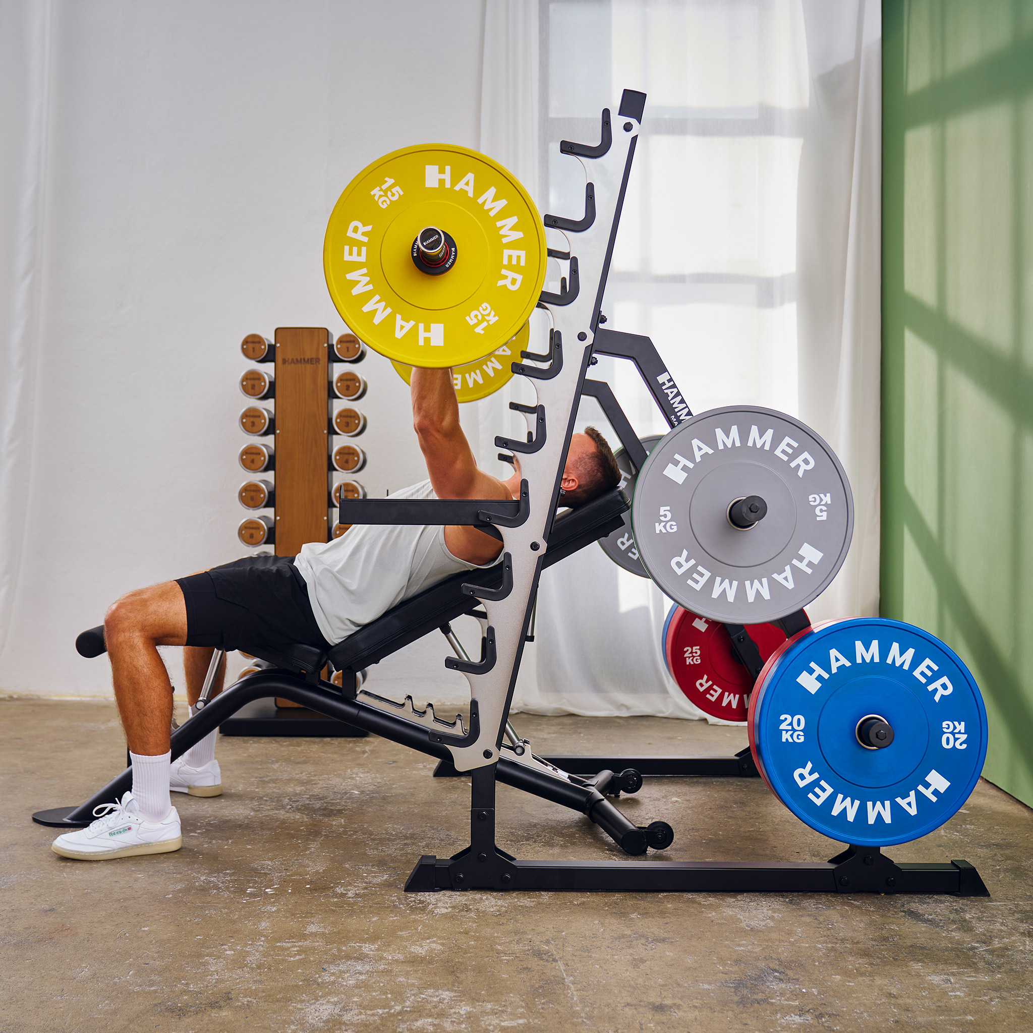 Mann Bankdrücken mit gelben Gewichten. Fitnessgeräte umfassen Hantelständer, Bank und verschiedene farbige Gewichte.
