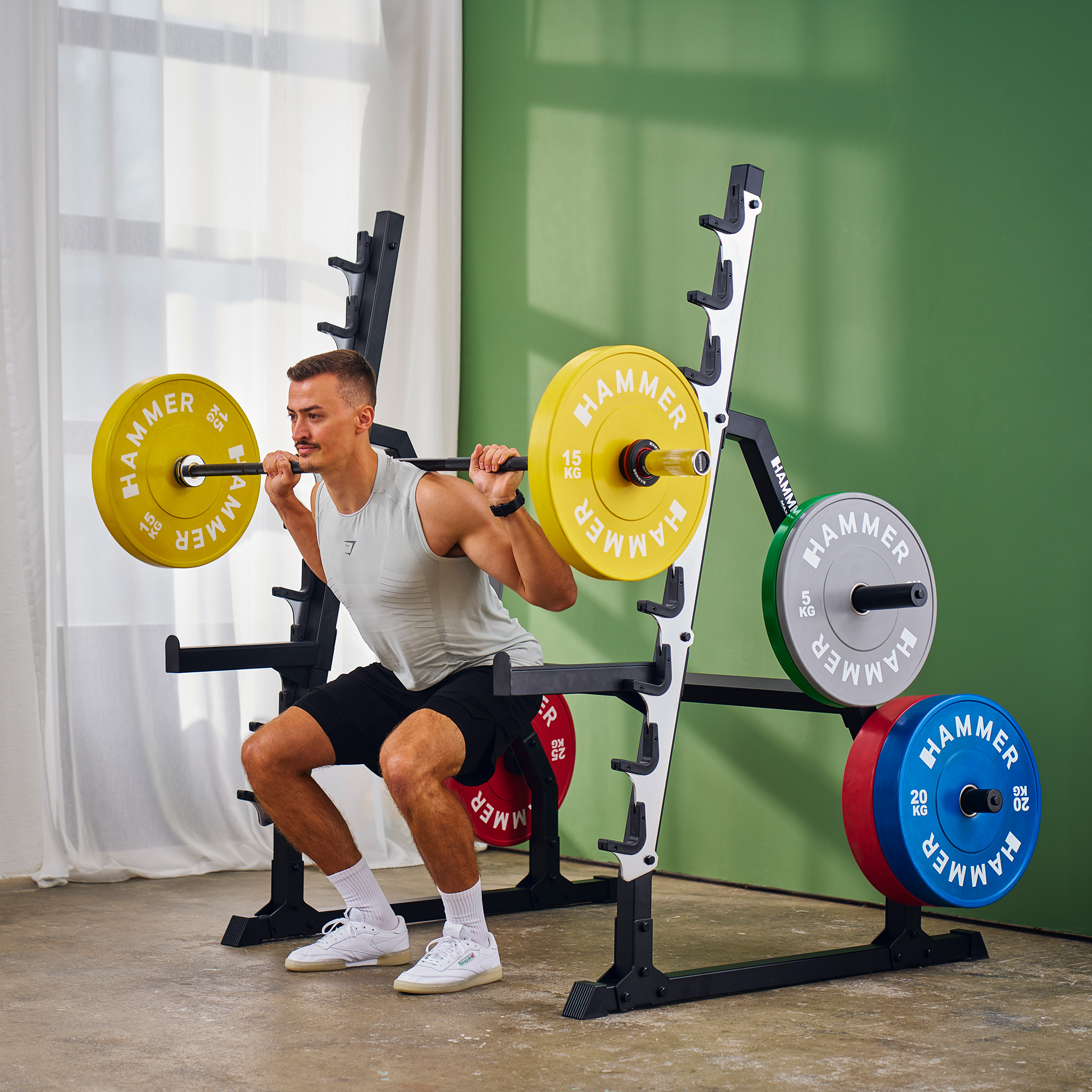 Mann macht Kniebeugen mit einer Langhantel im Fitnessstudio. Gewichte auf dem Ständer. Gelbe, rote, grüne und blaue Gewichte sind sichtbar.