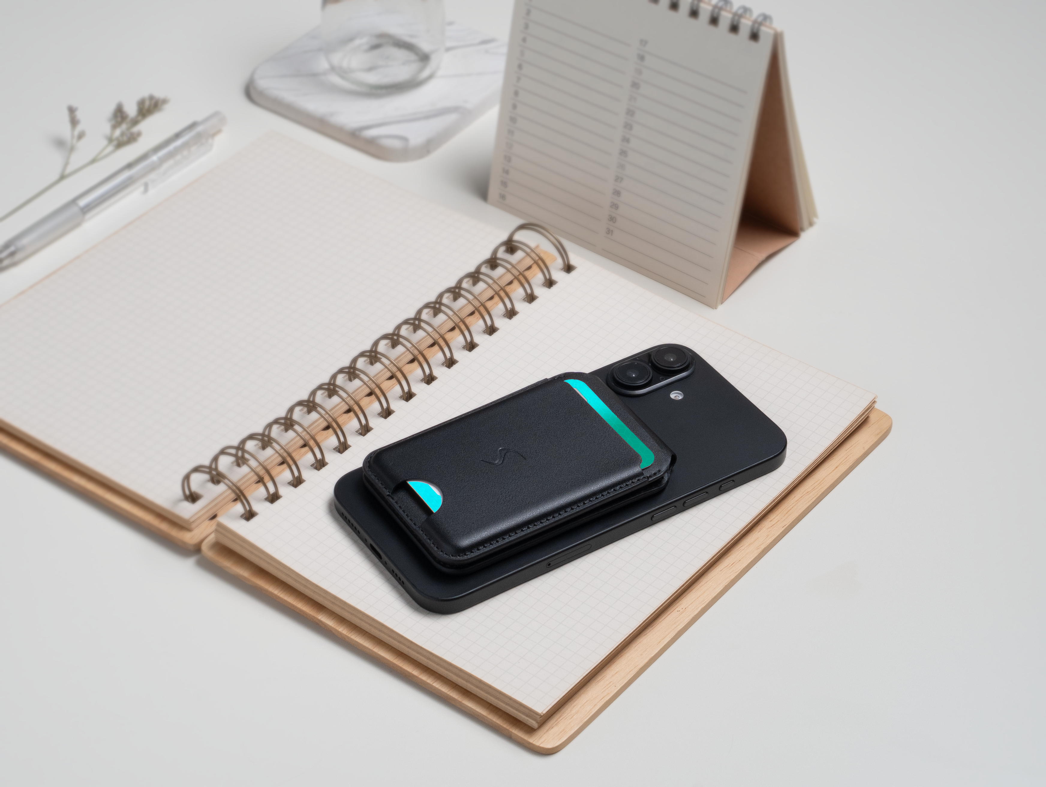 Schwarzes Smartphone, Brieftasche und Notizbuch auf weißer Oberfläche. Kalender und Stift sind ebenfalls sichtbar.