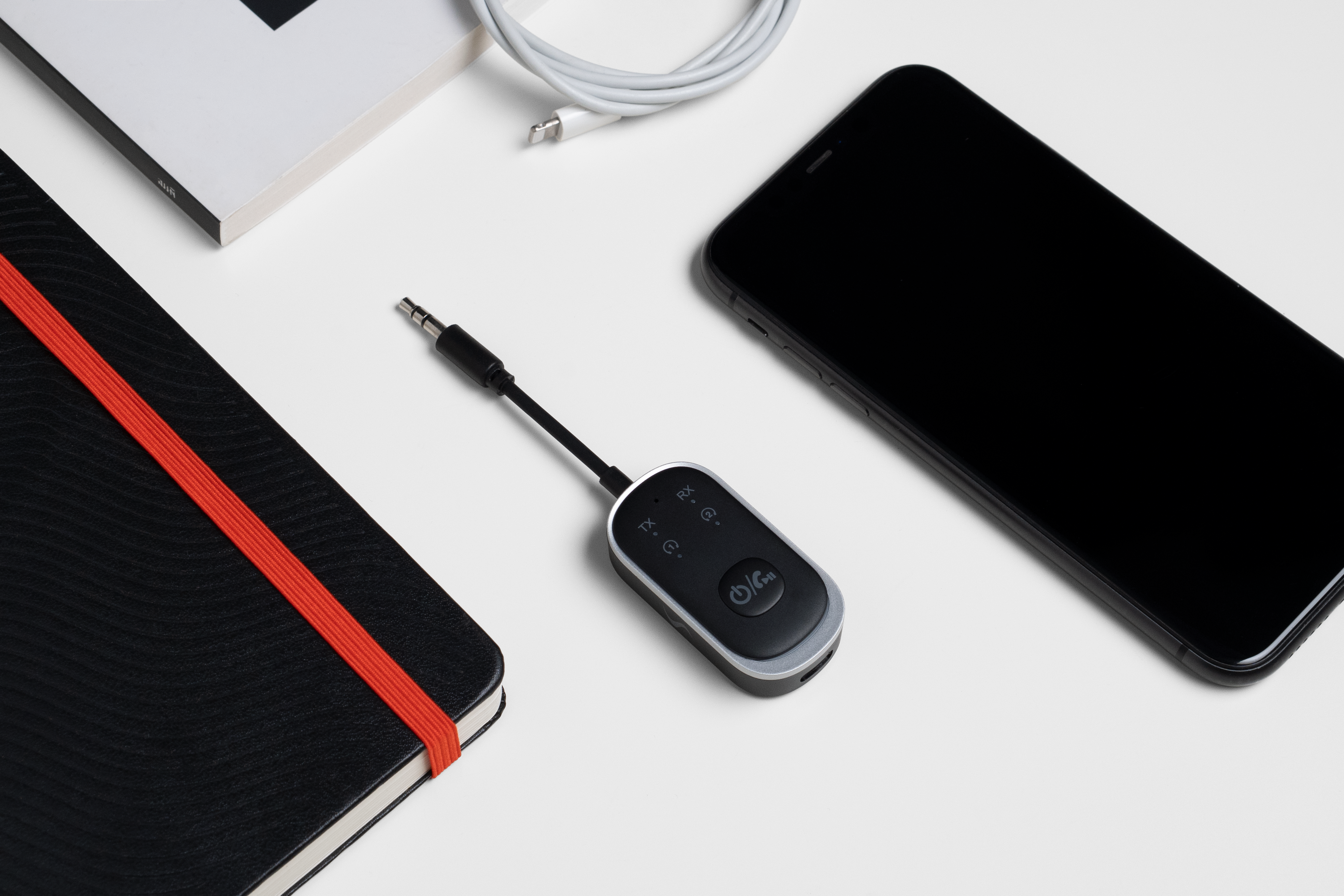 Ein schwarzes Smartphone, ein schwarzes Notizbuch mit rotem Band, ein Kabel und ein schwarzer Audioempfänger auf weißer Oberfläche.