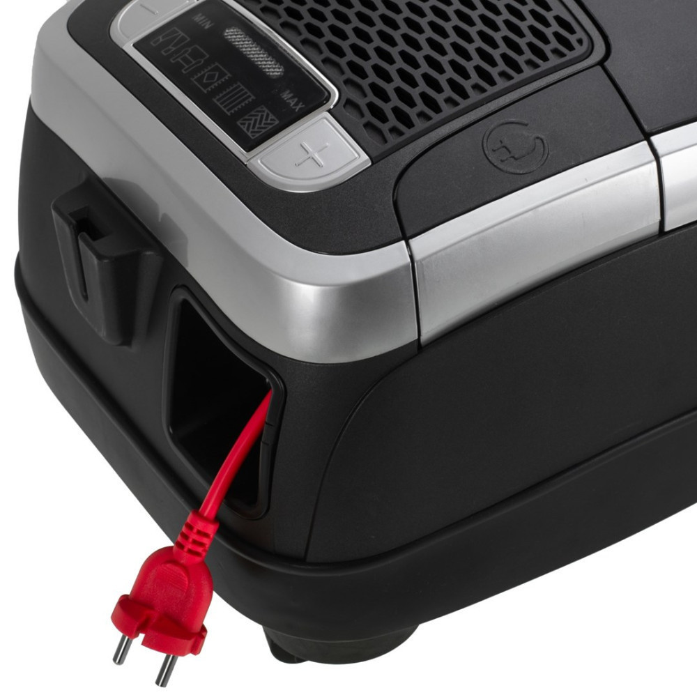 Zbliżenie odkurzacza. Czarny korpus ze srebrnym wykończeniem. Czerwony przewód zasilający jest włożony.