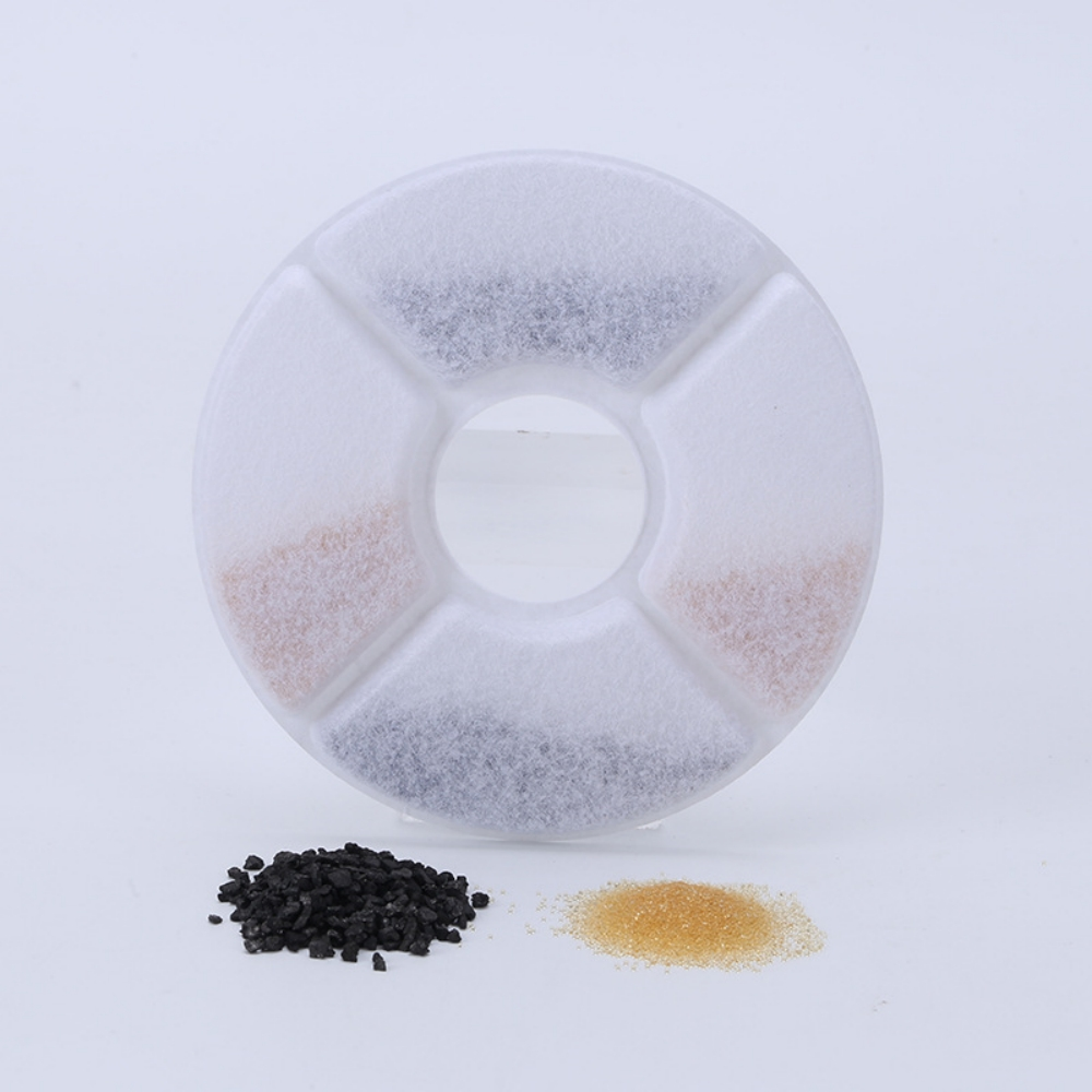 Ein weißer, kreisförmiger Katzenwasserfilter mit vier Abschnitten, die schwarze und braune Granulate enthalten. Zwei Granulathaufen darunter.