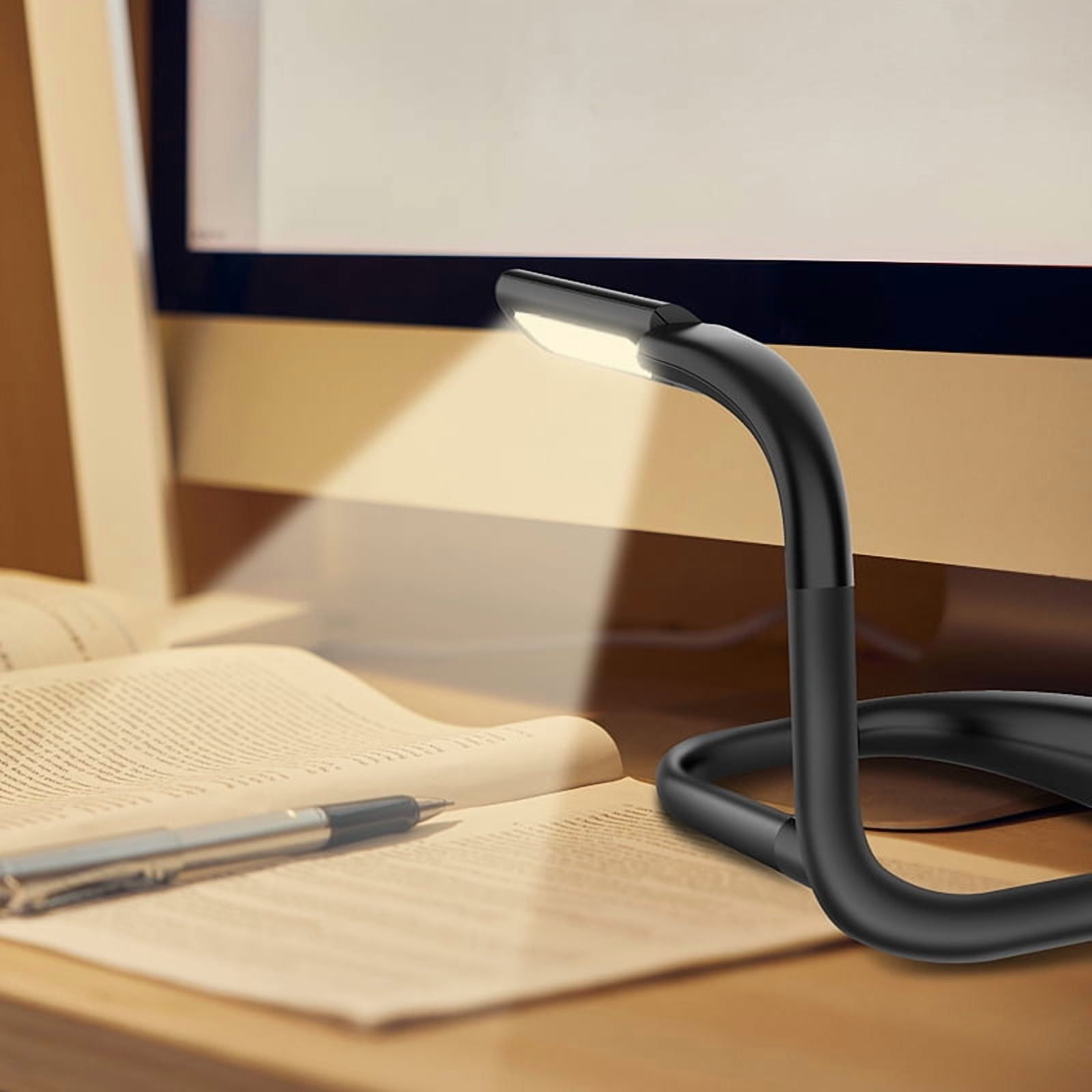 Eine schwarze Schreibtischlampe beleuchtet ein offenes Buch. Ein Stift liegt auf der Seite. Ein Computerbildschirm ist im Hintergrund.