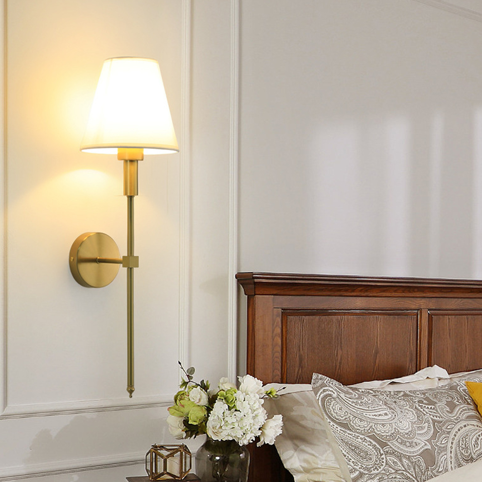 Schlafzimmer mit Wandlampe, Bett, Blumen und Kissen. Die Lampe ist golden. Das Bett ist aus Holz.