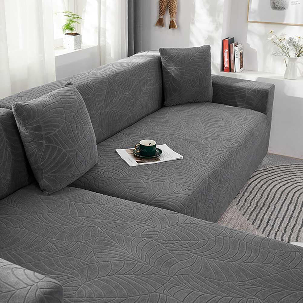 Graues Sofa mit Blättermuster, Kissen und einer Kaffeetasse auf Papier. Im Hintergrund ein Fenster und ein Regal.