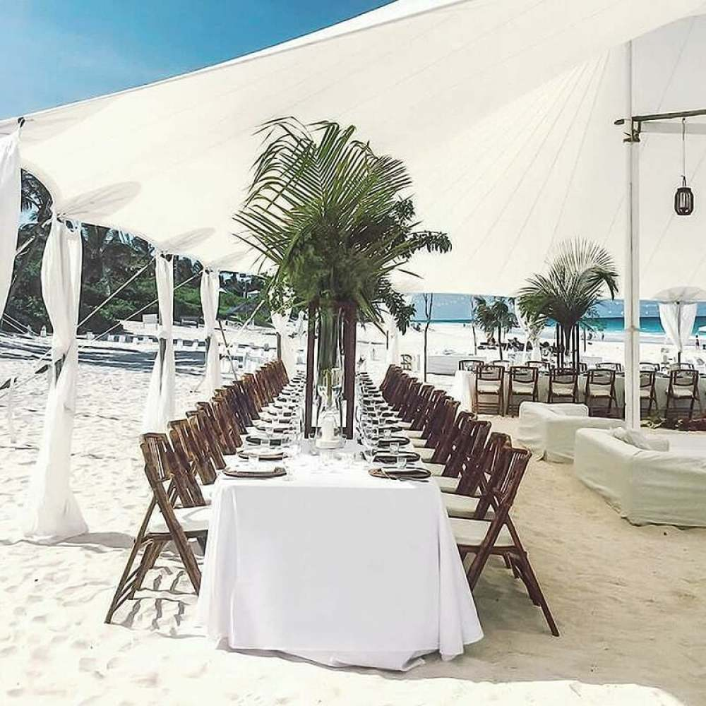 Strandhochzeit. Langer Tisch mit weißem Tuch, Stühlen, Palmen und Baldachin im Sand. Meer und Himmel im Hintergrund.