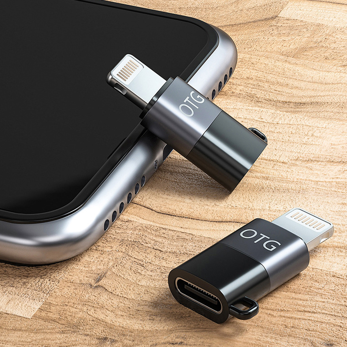 Zwei OTG-Adapter, einer an einem Telefon angeschlossen. Einer silberfarben, einer schwarz, auf einer Holzoberfläche. Adapter haben USB-C- und Lightning-Anschlüsse.