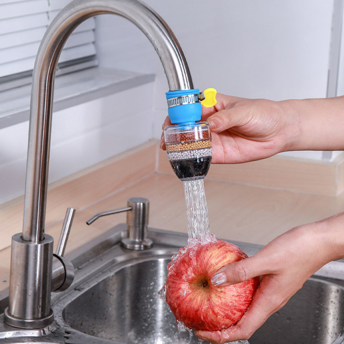 Ein Wasserhahn mit Filter besprüht einen roten Apfel, der von Händen in einem Küchenbecken gehalten wird. Der Filter ist blau und transparent.