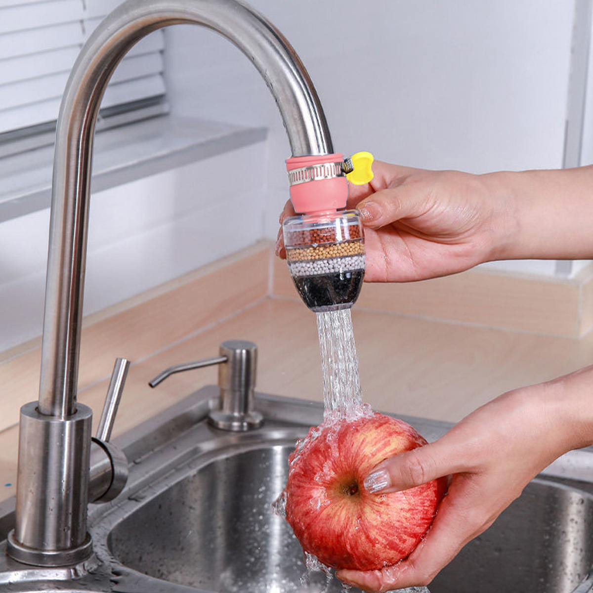 Eine Person wäscht einen Apfel unter einem Wasserhahn mit Wasserfilter. Der Apfel ist rot, das Wasser fließt in ein Edelstahlbecken.
