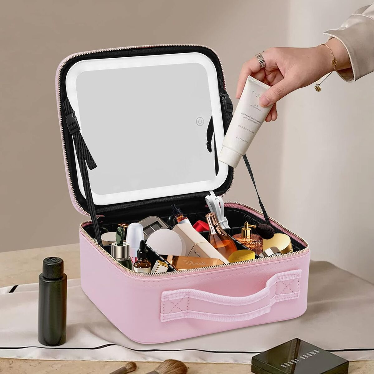 Un estuche de maquillaje rosa abierto con espejo y cosméticos. Una mano sostiene un tubo de crema. Otros artículos son visibles en la mesa.