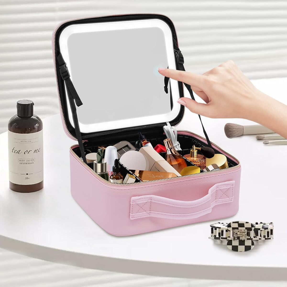 Un estuche de maquillaje rosa está abierto sobre una superficie blanca, revelando cosméticos y un espejo. Una mano toca el espejo.