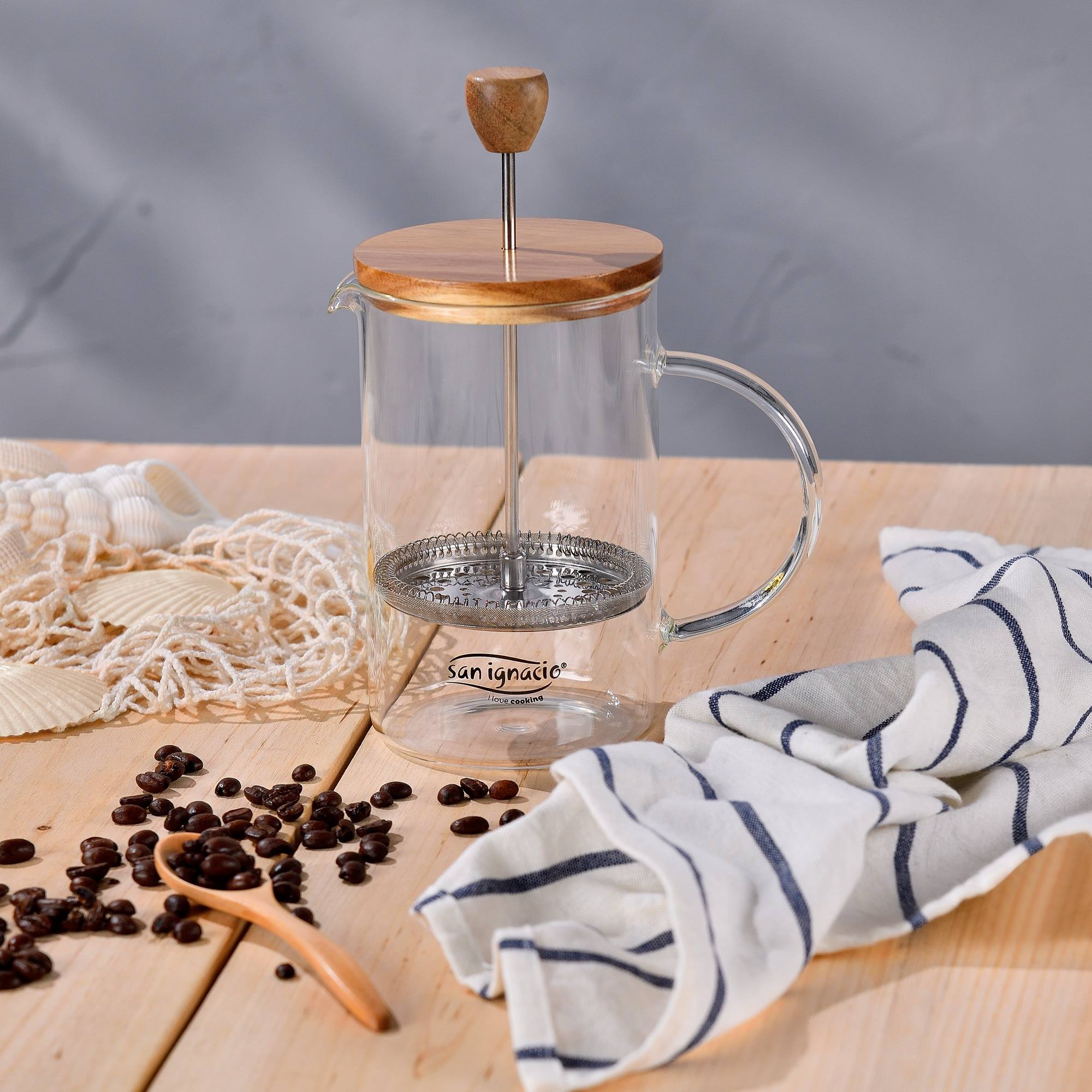 Prensa francesa sobre una mesa de madera. Granos de café y una cuchara cerca. Una toalla blanca y azul a la derecha.