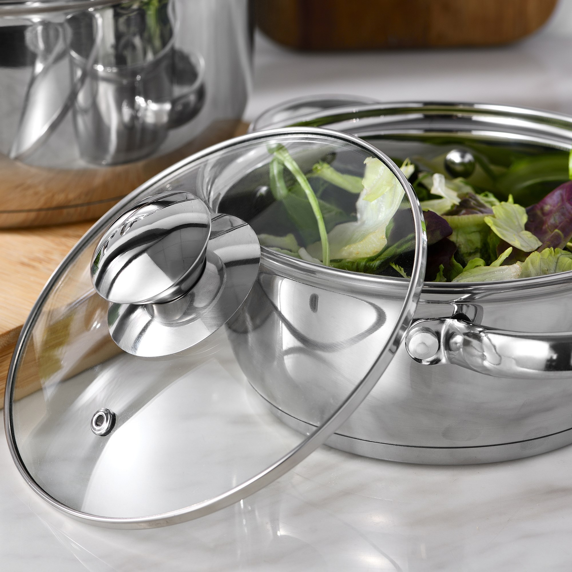 Una olla de acero inoxidable con tapa de cristal. Dentro hay una ensalada. Otra olla en el fondo sobre una tabla de madera.