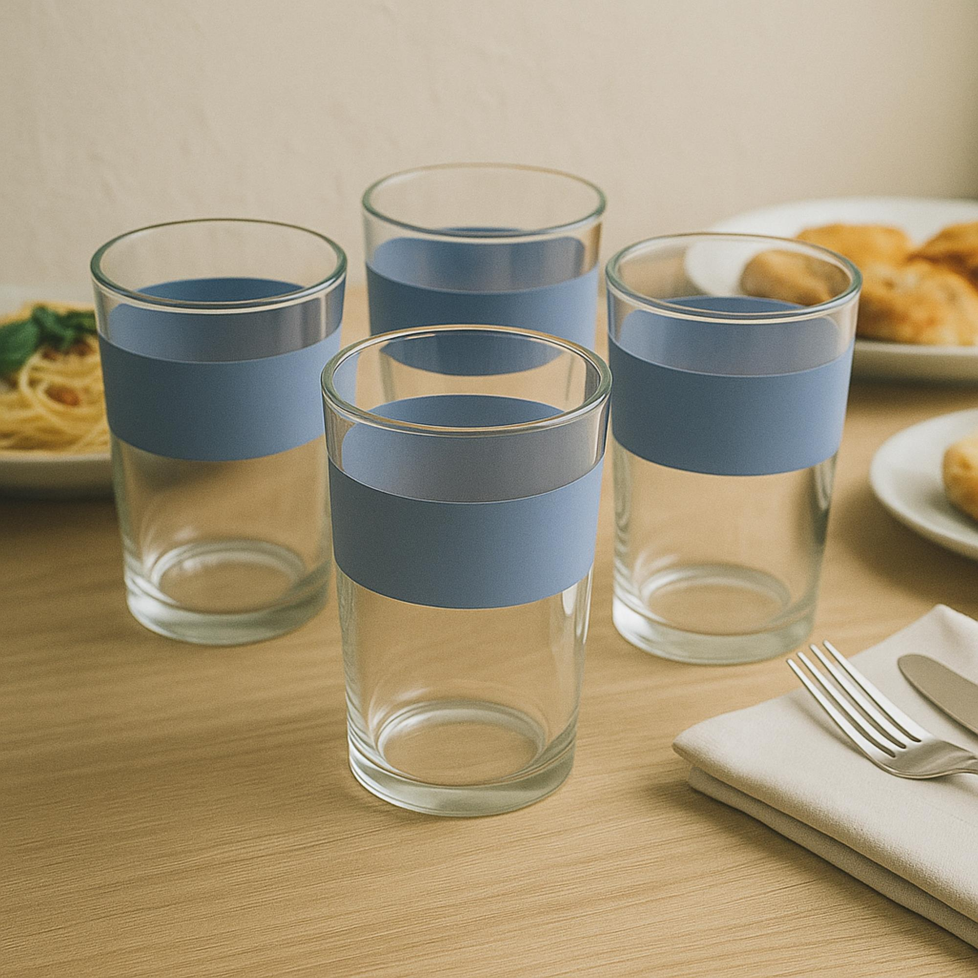 Cuatro vasos transparentes con bandas azules sobre una mesa de madera. Platos con comida y cubiertos al fondo.