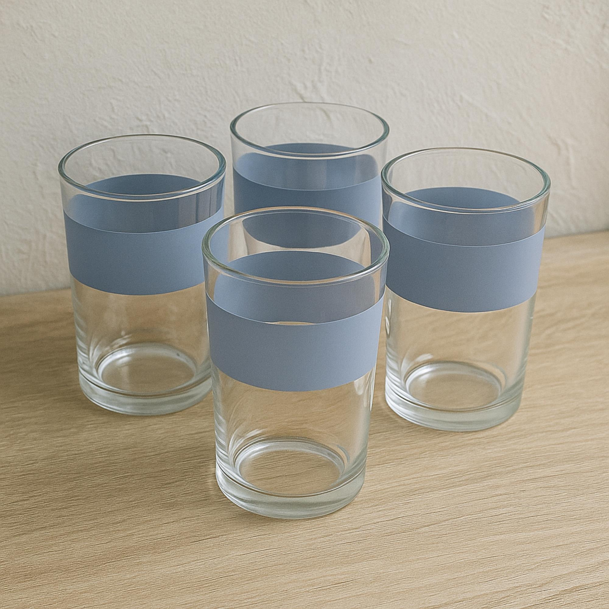 Cuatro vasos de vidrio transparente con bandas azules sobre una superficie de madera clara, contra una pared blanca texturizada.