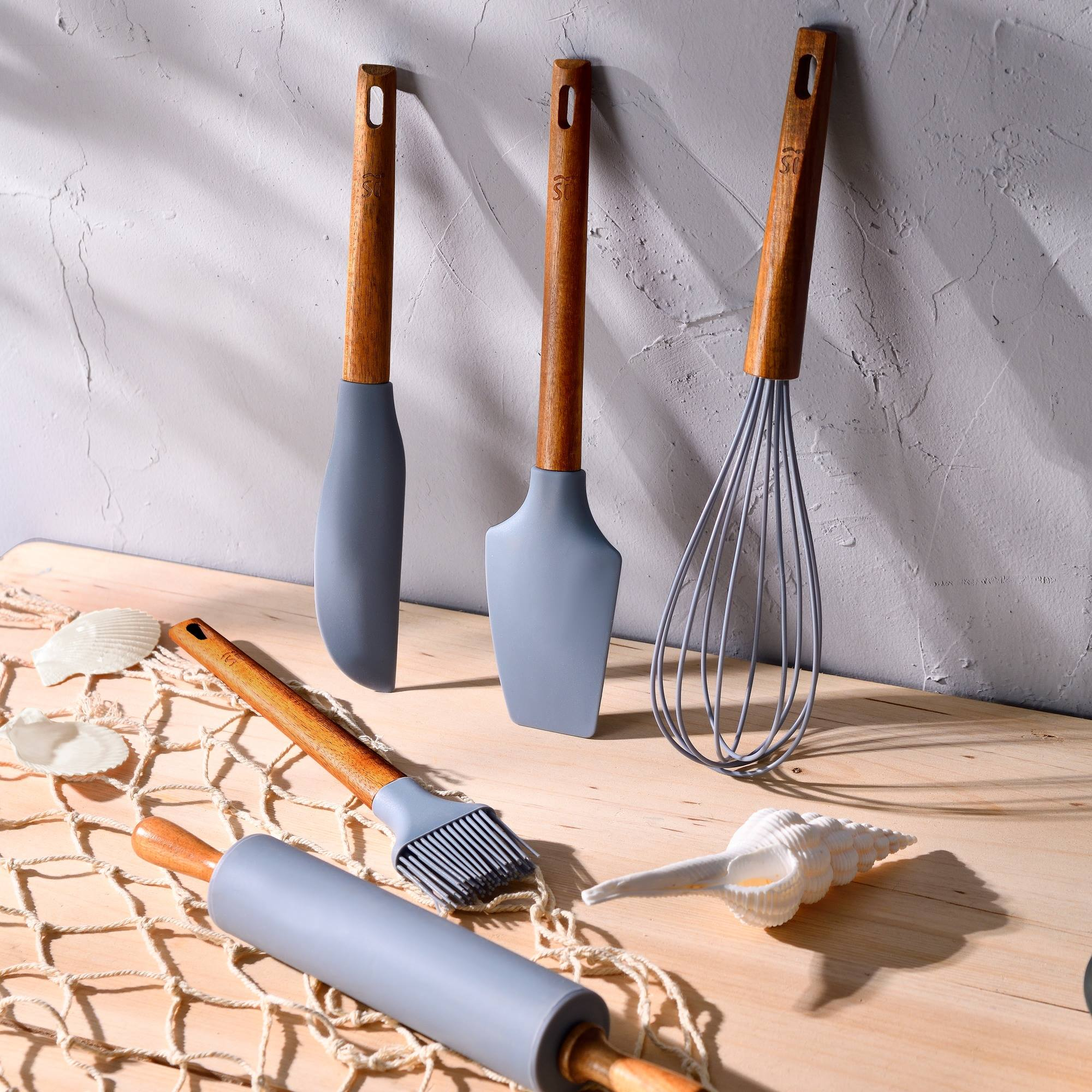 Utensilios de cocina sobre una superficie de madera. Incluye espátula, batidor, pincel y rodillo con conchas.