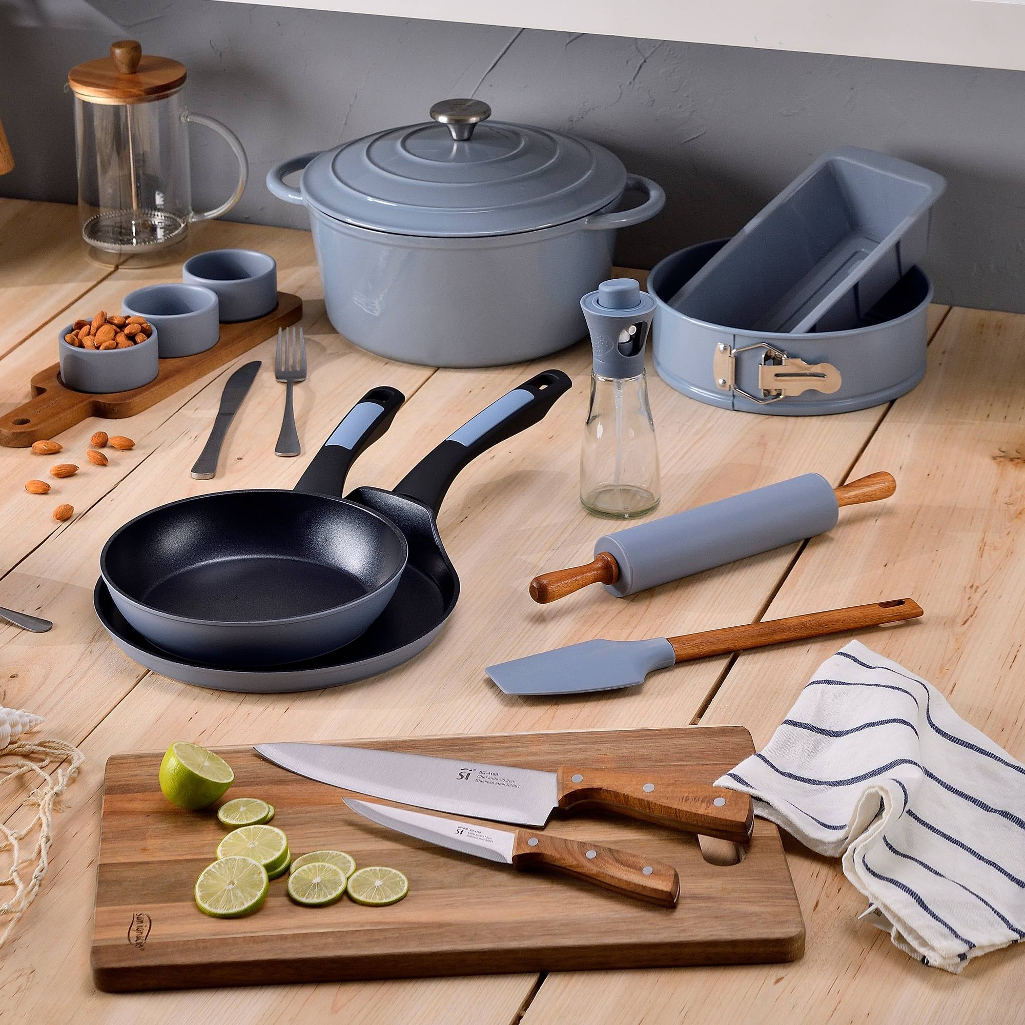 Utensilios de cocina sobre superficie de madera: sartenes, ollas, cuchillos, rodillo y tabla de cortar con limas, almendras y tela.