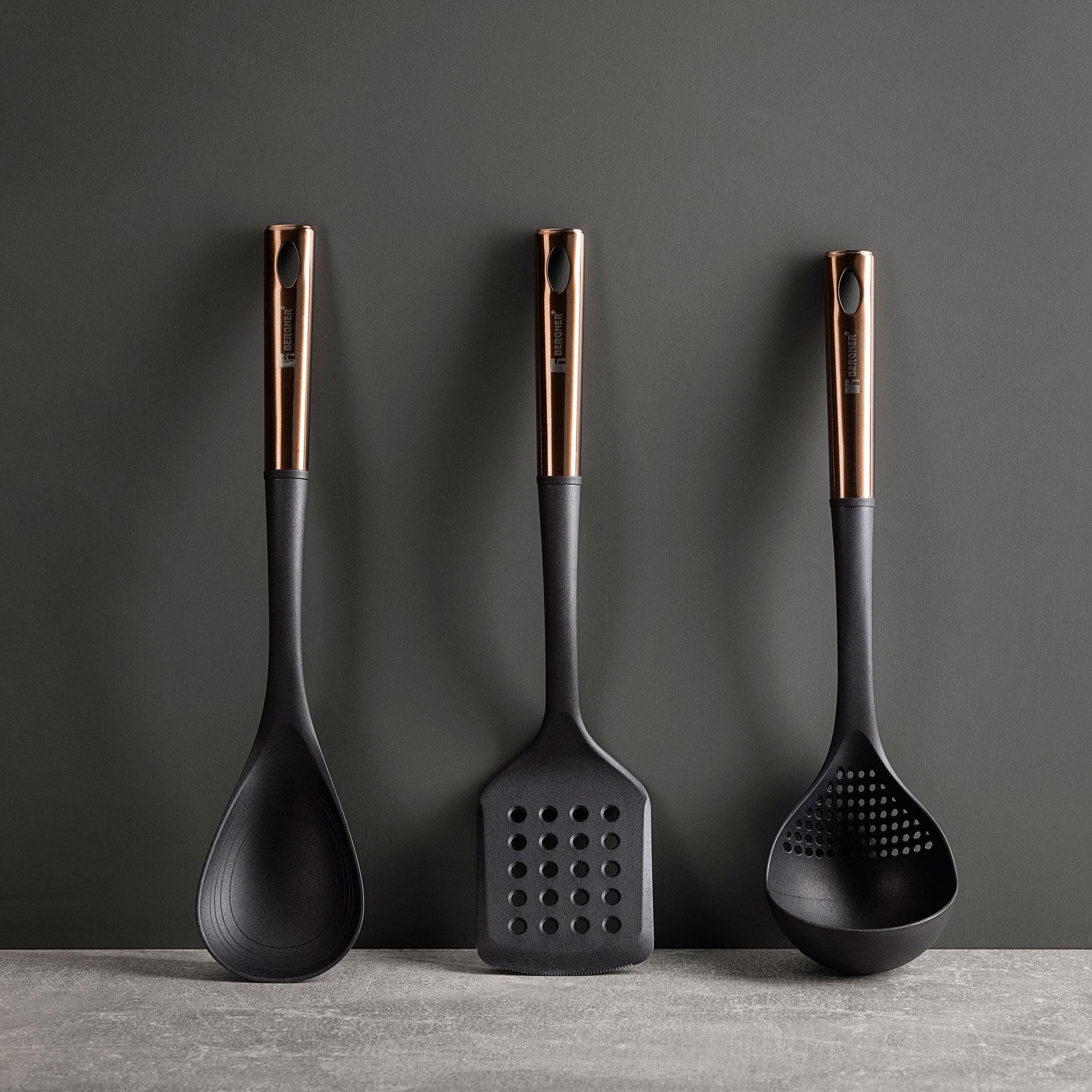 Tres utensilios de cocina negros con mangos dorados están dispuestos verticalmente sobre un fondo gris.