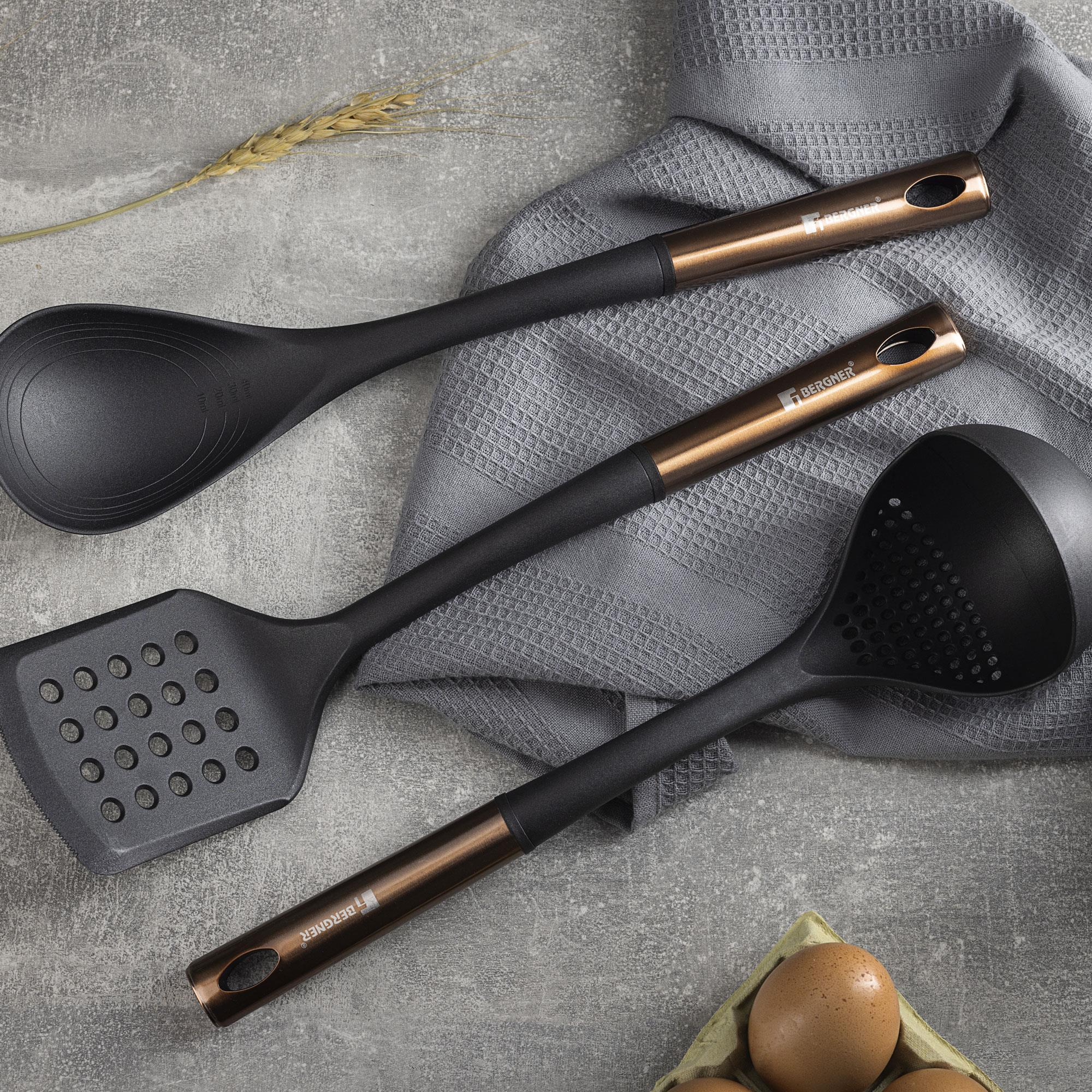 Utensilios de cocina negros, incluyendo cuchara, espátula y cucharón ranurado, sobre superficie gris, toalla y huevos.