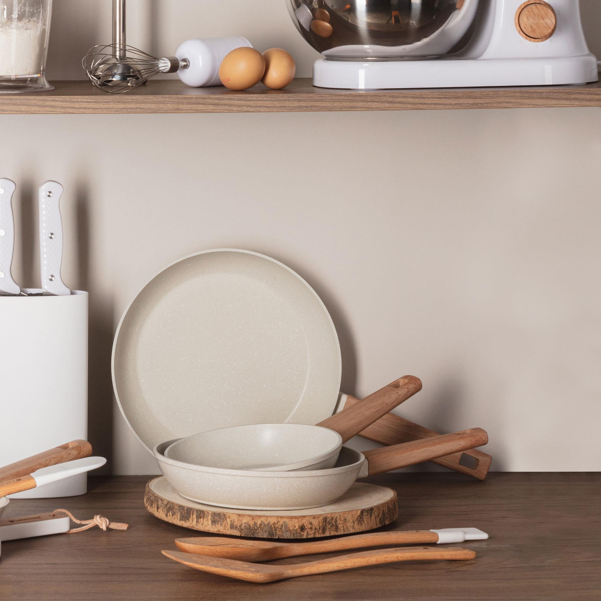 Configuración de cocina con sartenes, utensilios, batidora y huevos sobre una superficie y estante de madera.