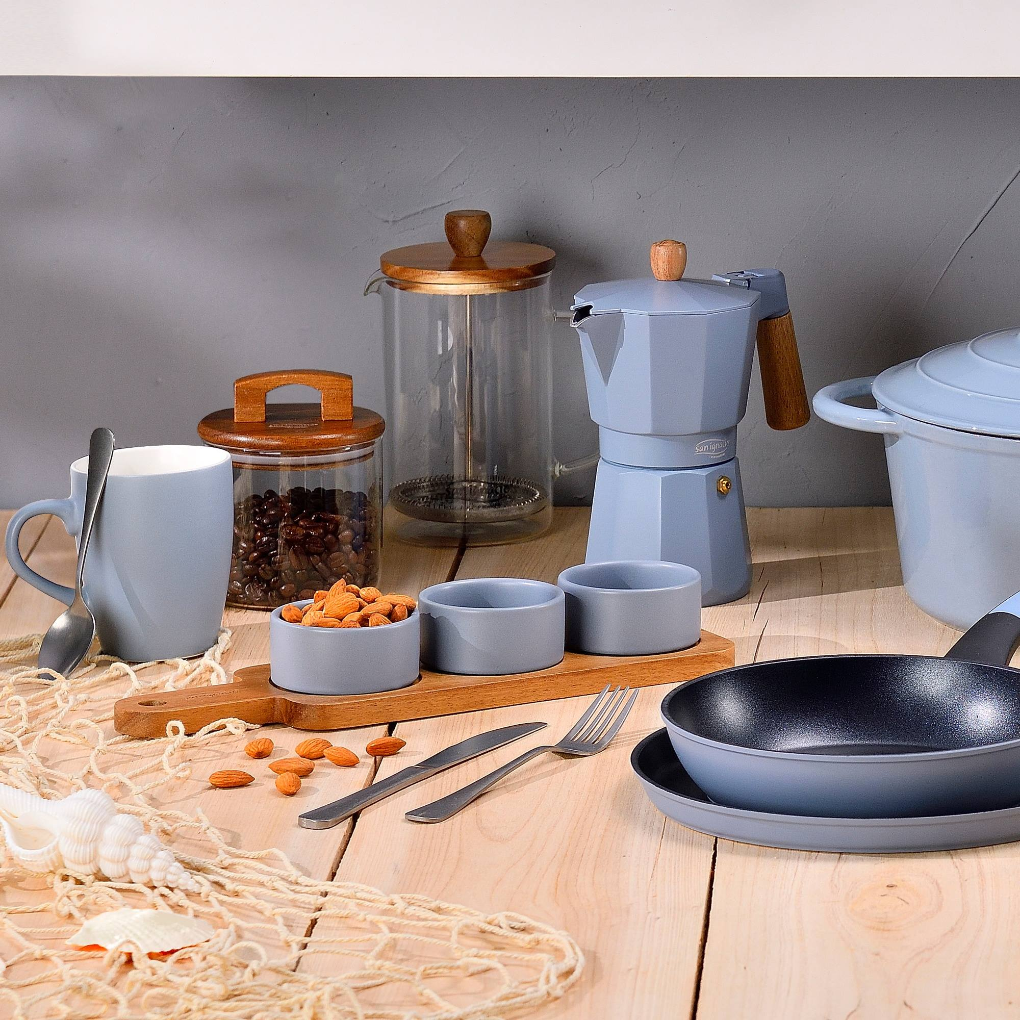 Configuración de cocina con cafetera, ollas, taza, cuencos con almendras, utensilios y una concha sobre una mesa de madera.