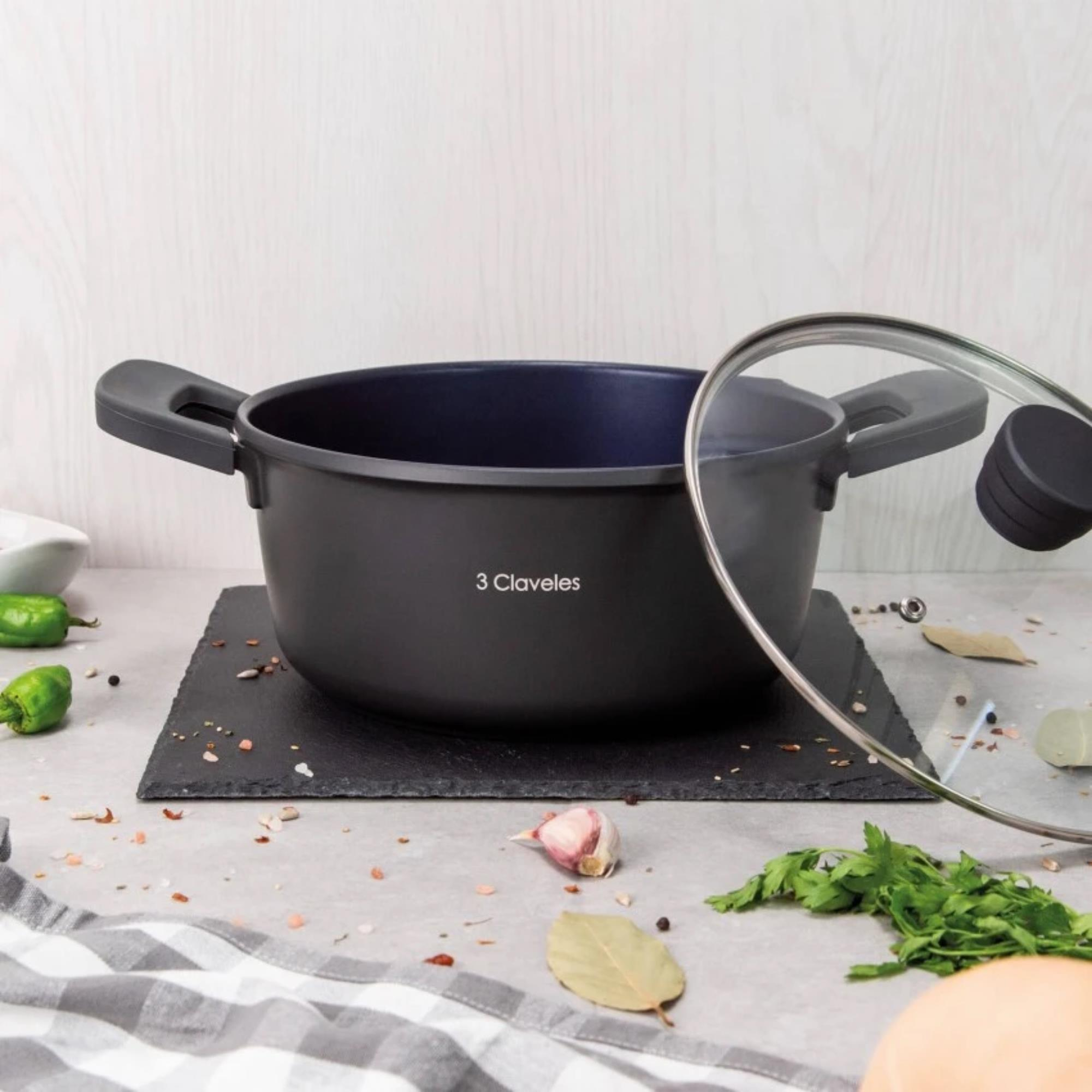 Una olla negra con tapa de cristal, sobre una placa oscura, rodeada de ingredientes como pimientos, ajo y hierbas.