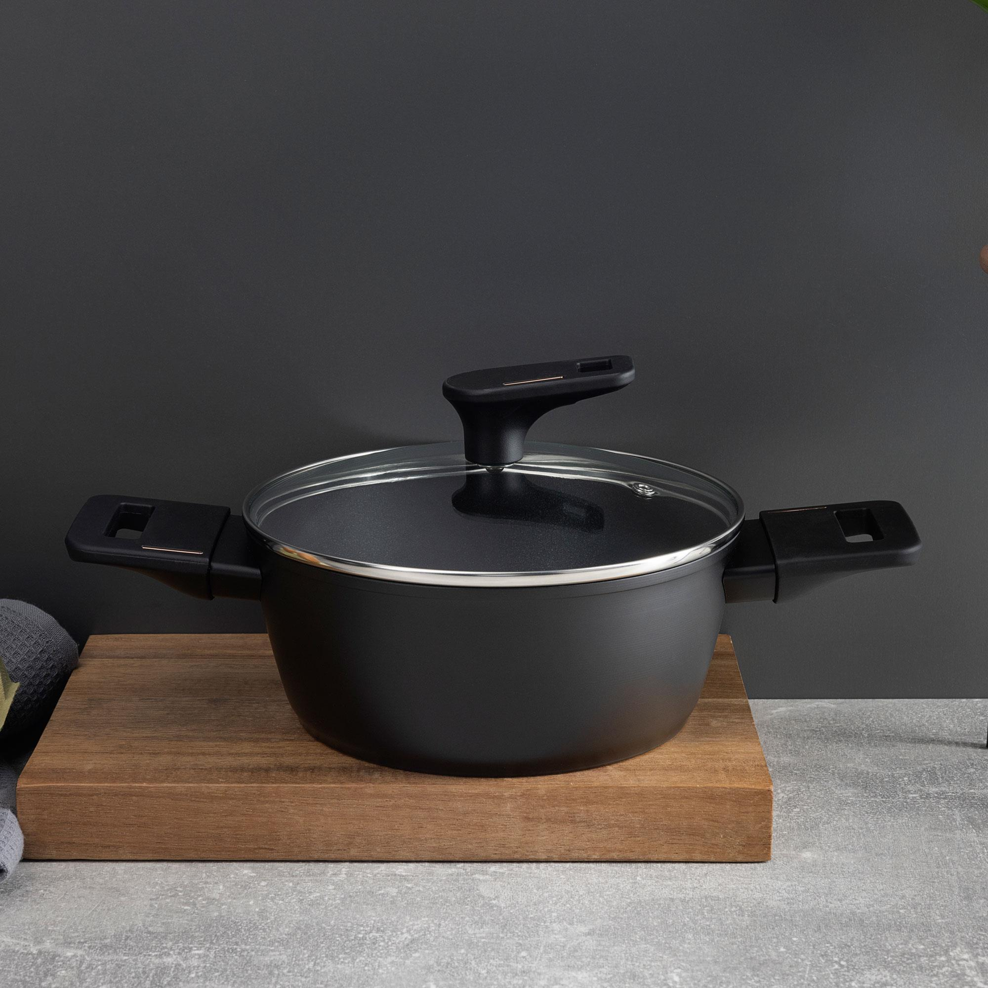 Una olla negra con tapa de cristal sobre una tabla de madera. El fondo es una pared gris oscuro y una superficie de hormigón.
