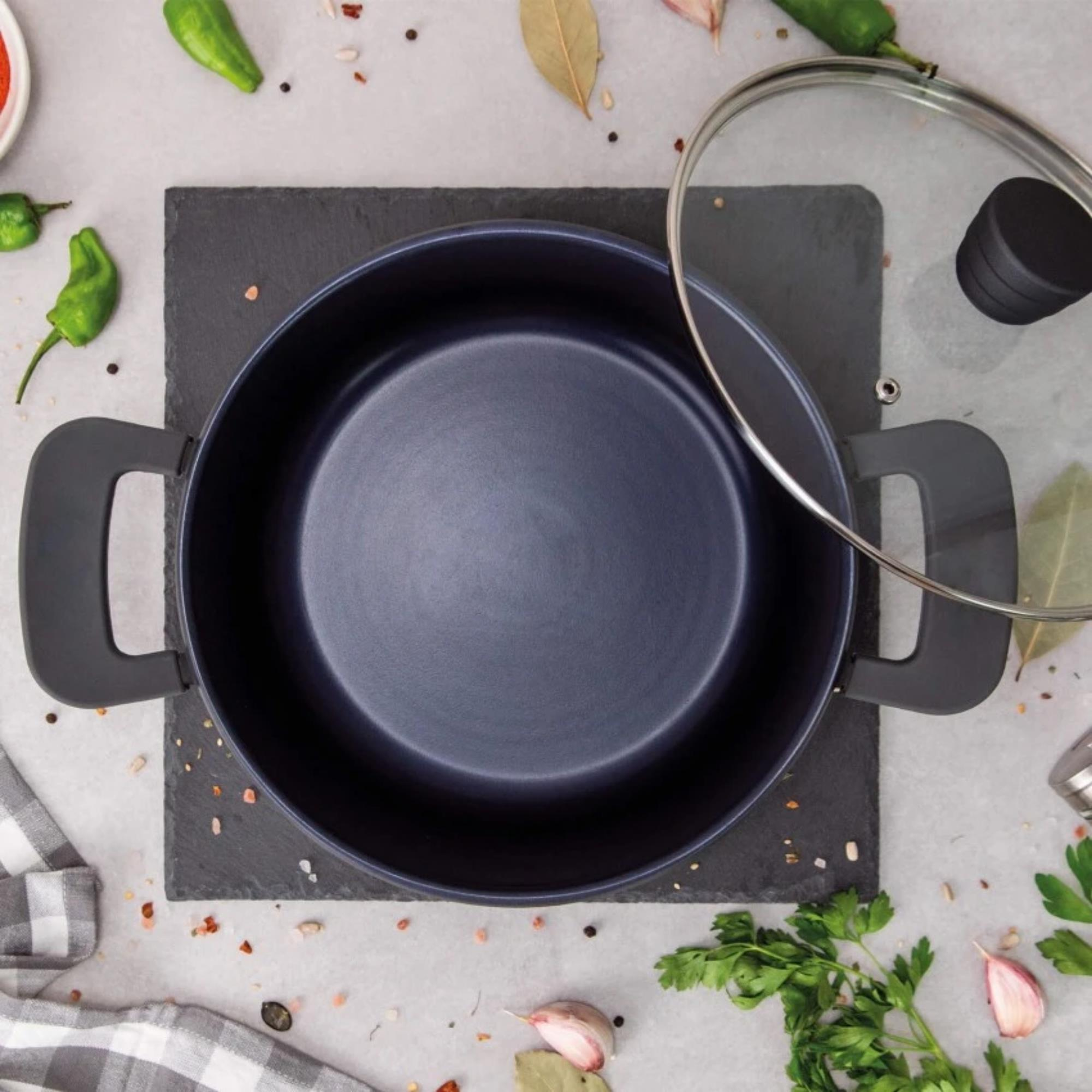 Una olla azul oscura sobre una pizarra negra. Ingredientes como pimientos, perejil y ajo están esparcidos.