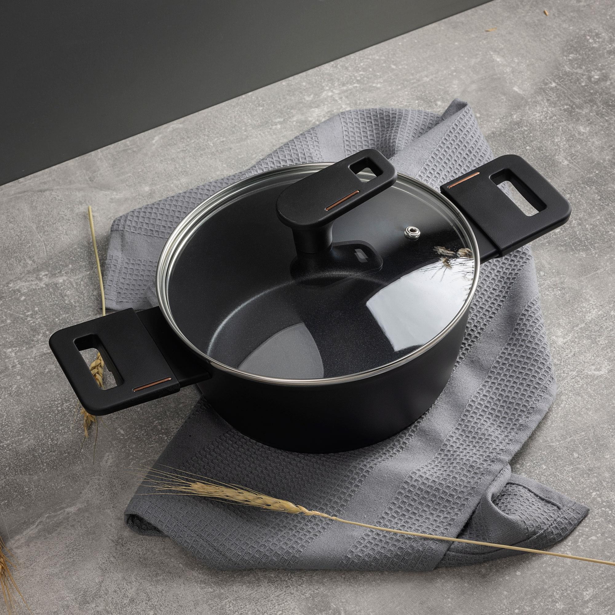 Olla negra con tapa de cristal, sobre una toalla gris, con espigas de trigo. La olla tiene asas negras. El fondo es gris.