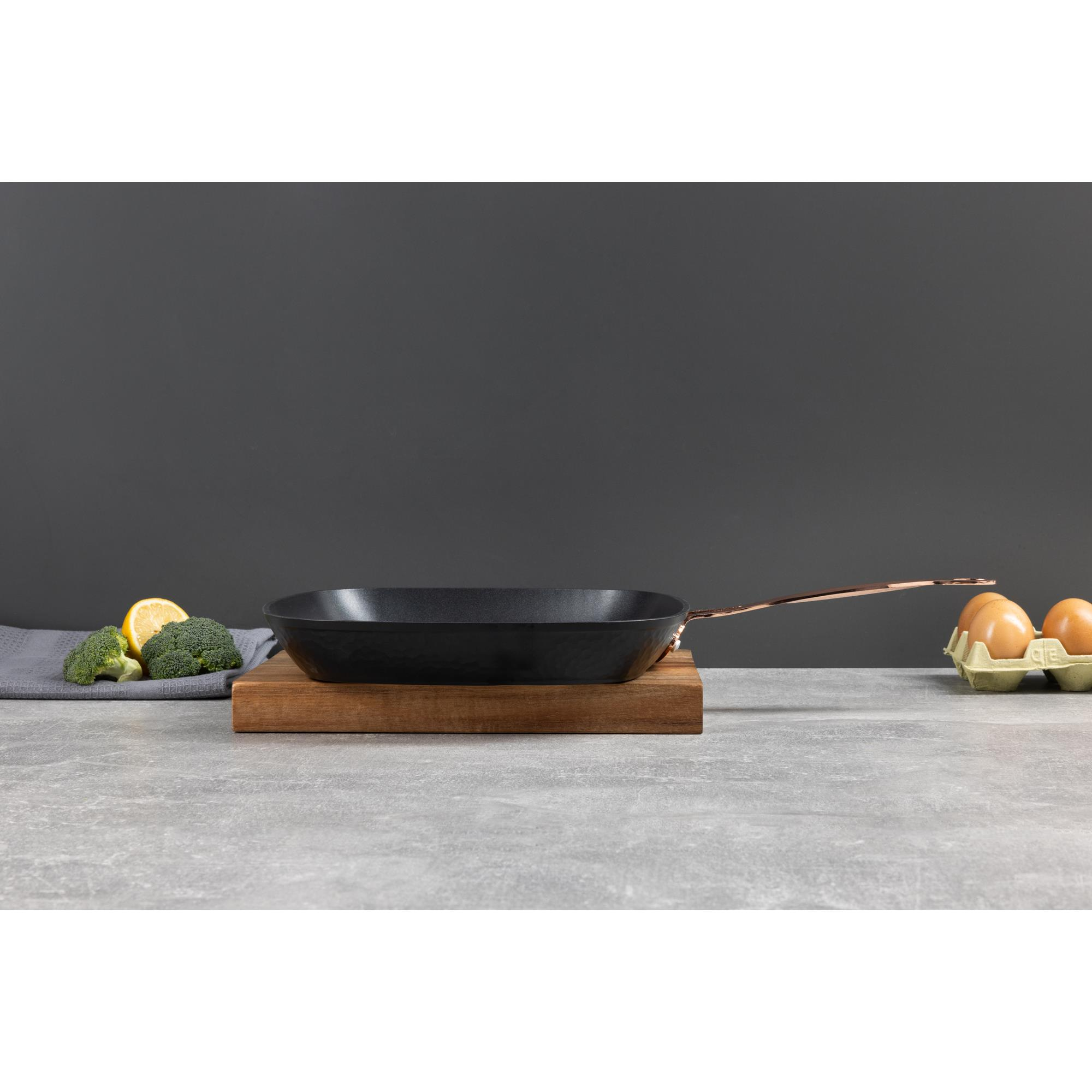 Una sartén negra sobre una tabla de madera con brócoli, limón y huevos sobre un fondo gris.