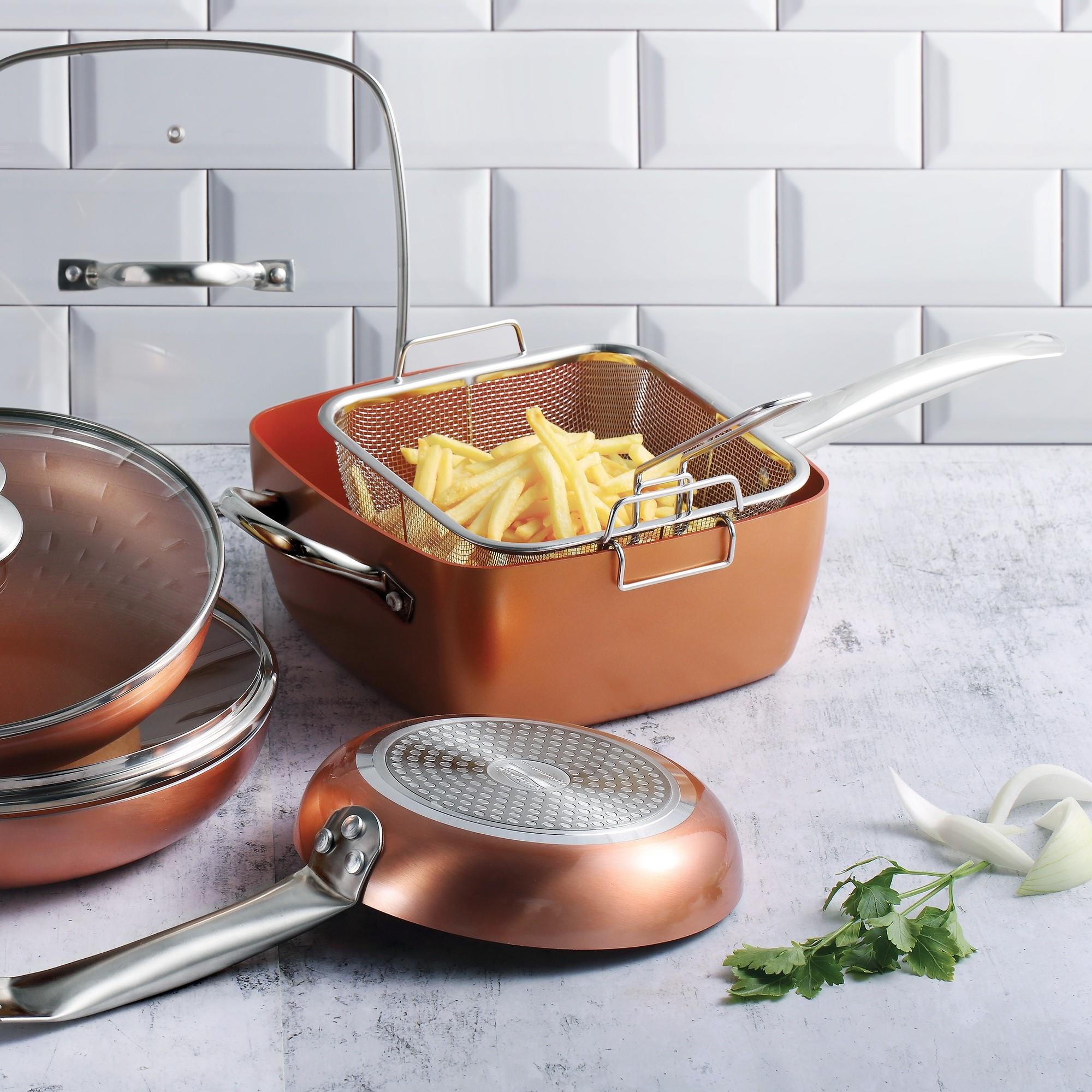 Juego de utensilios de cocina de cobre con papas fritas en una cesta. Incluye sartenes y tapas, sobre una superficie texturizada, fondo de pared de ladrillos blancos.