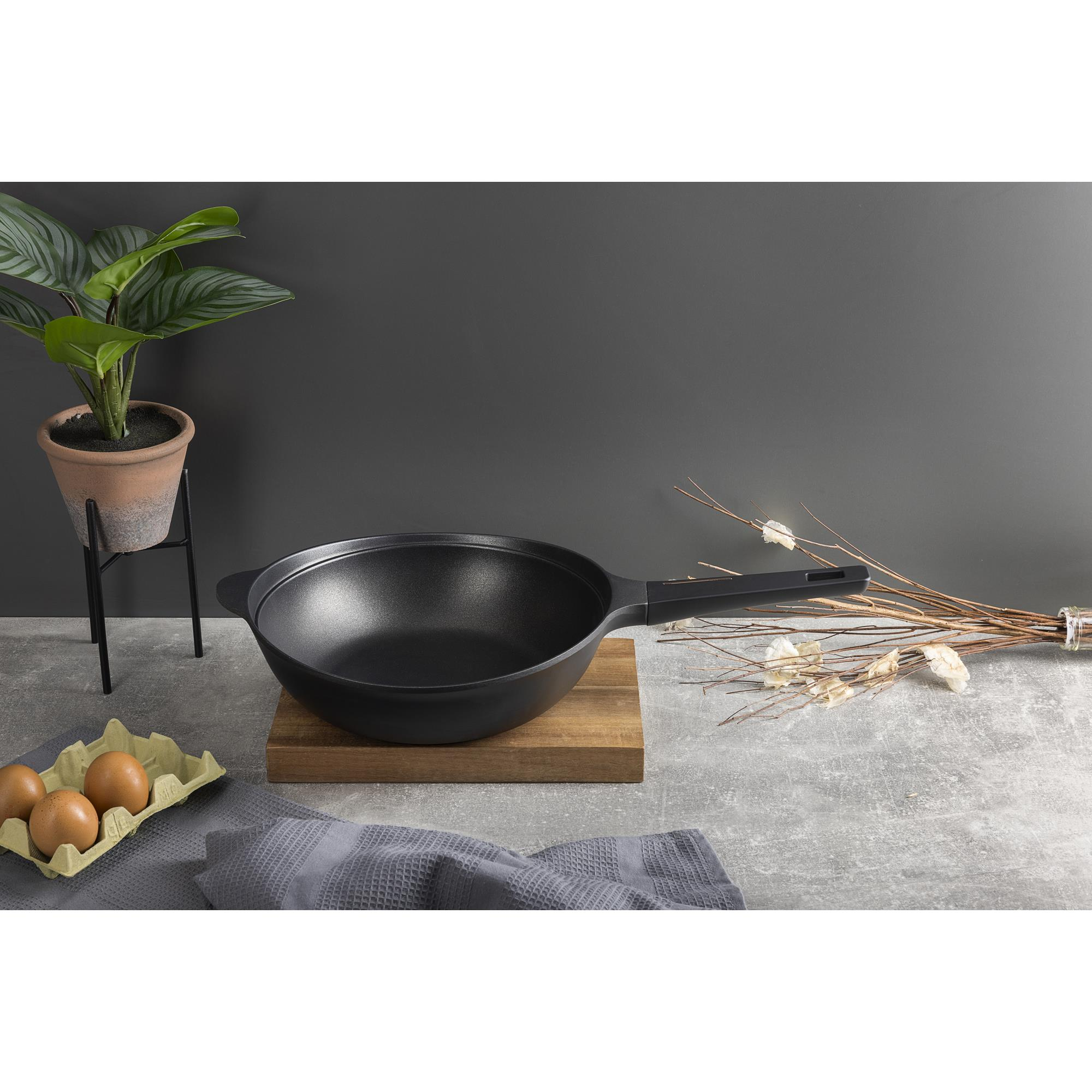 Wok negro sobre tabla de madera, planta en maceta, huevos en cartón y un paño gris sobre superficie gris.