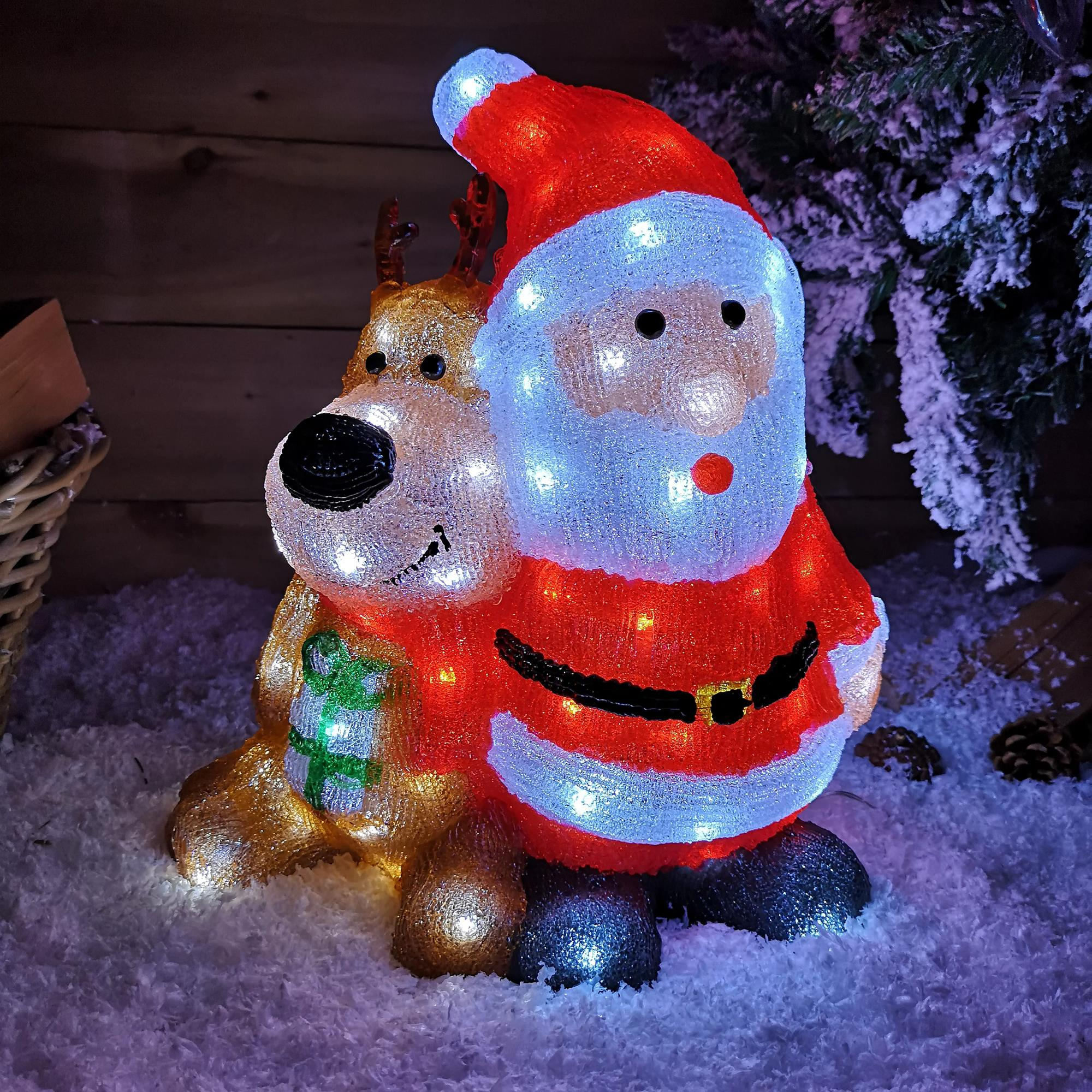 Decoración iluminada de Papá Noel y reno en la nieve. Papá Noel tiene un traje y gorro rojos, y el reno es dorado.