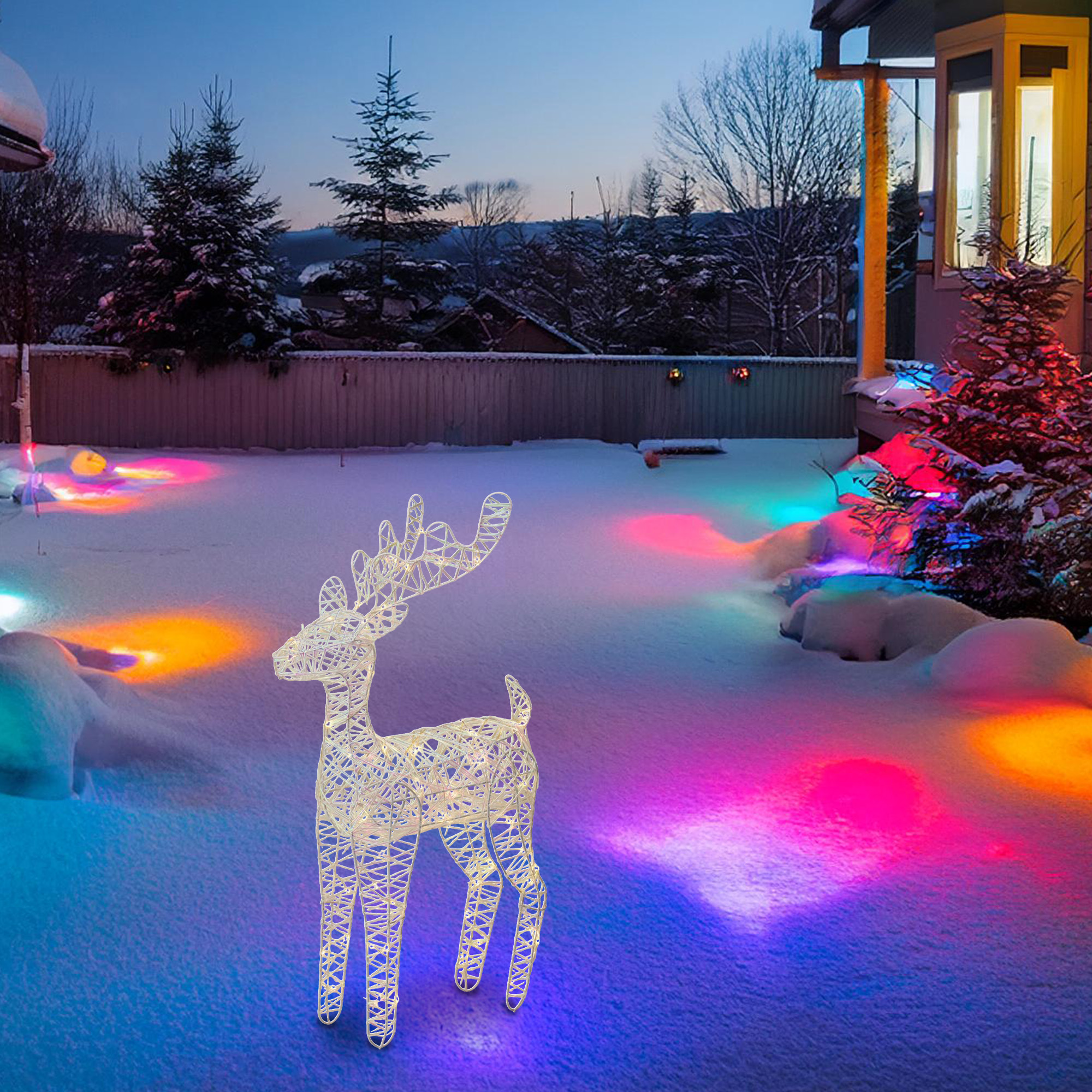 Un reno iluminado en la nieve, con luces de colores. Árboles y una casa al fondo. Es de noche.
