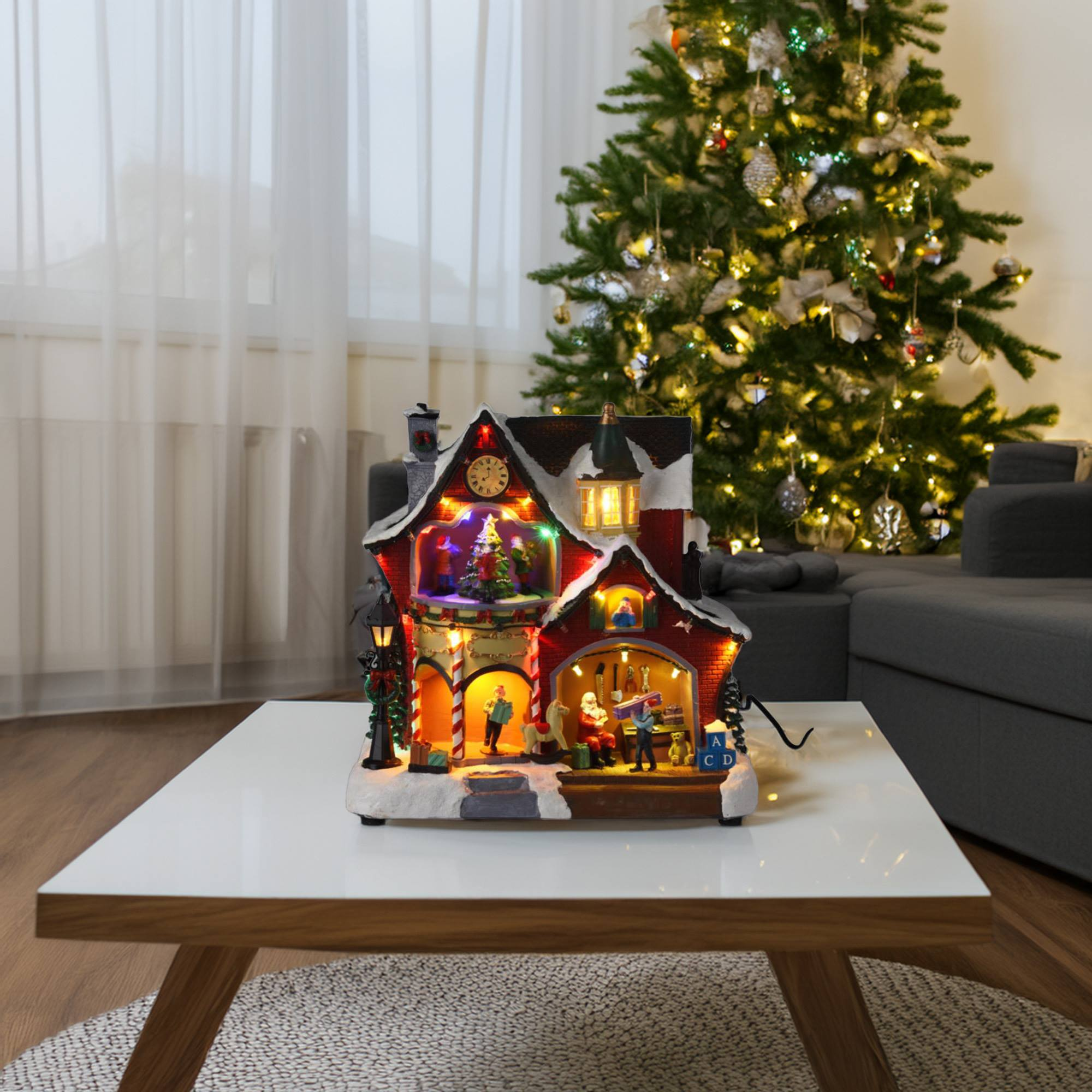Una escena de un pueblo navideño en una mesa blanca. Un árbol iluminado está al fondo. Papá Noel y otros están dentro de un edificio.