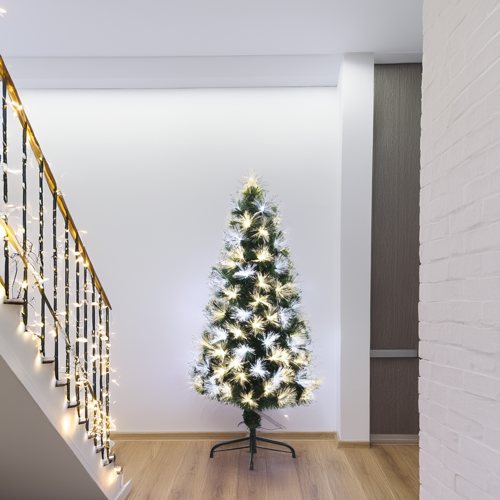 Un árbol de Navidad iluminado está en un pasillo. A la izquierda hay una escalera con luces. A la derecha hay una pared de ladrillos blancos.
